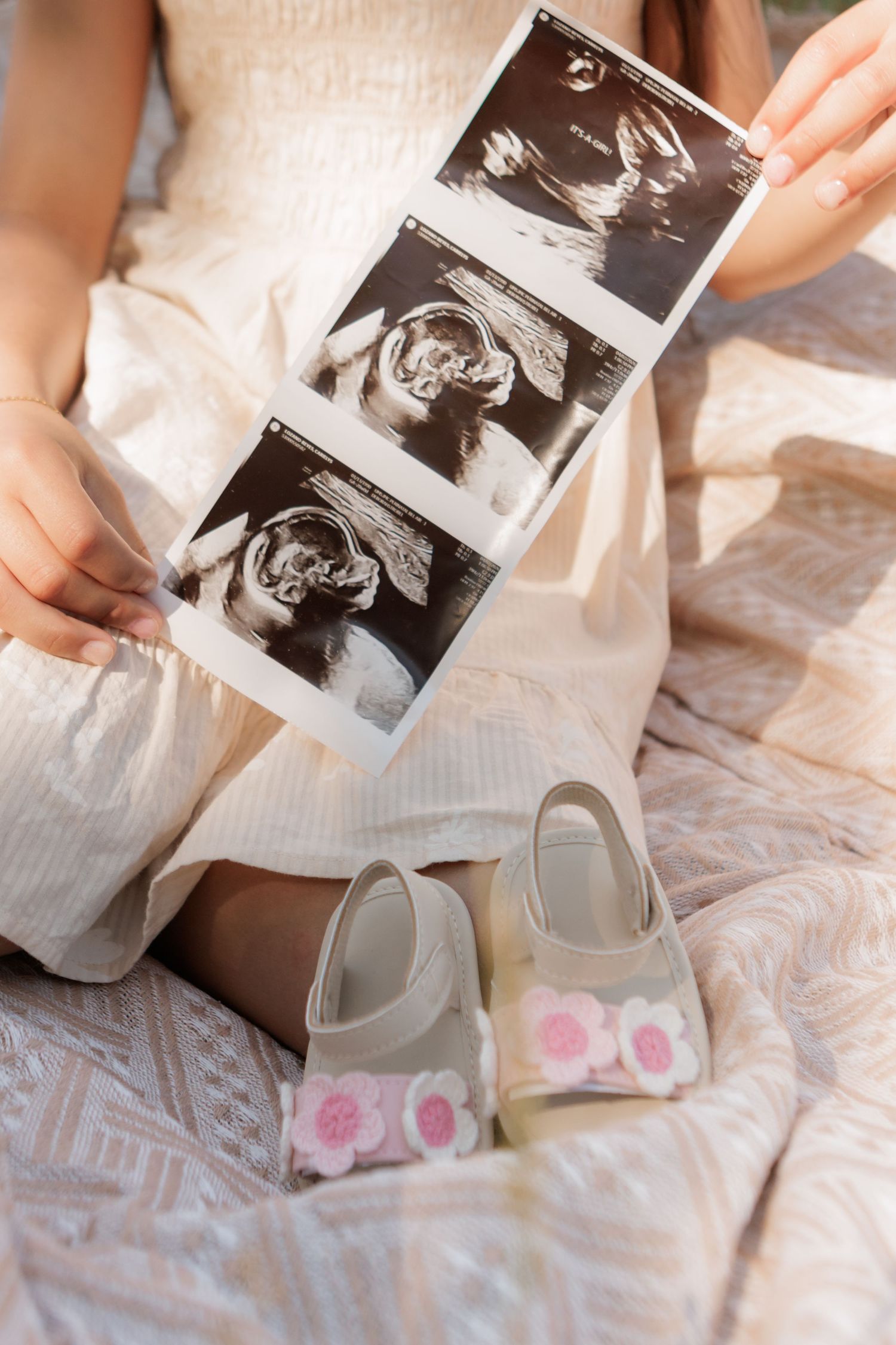 Woman in a cream dress sits on a blanket holding a three-photo ultrasound strip beside pink floral baby sandals, celebrating a pregnancy.