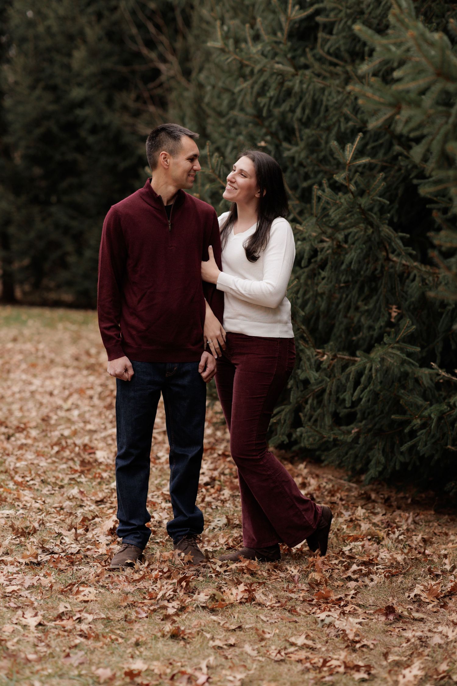 A couple in coordinating burgundy and white outfits poses playfully near evergreen trees.