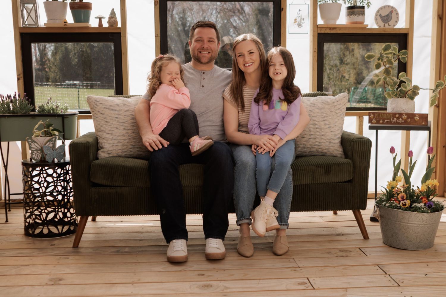 Family of four smiling together on a dark couch in a bright sunroom with plants and wooden floors.