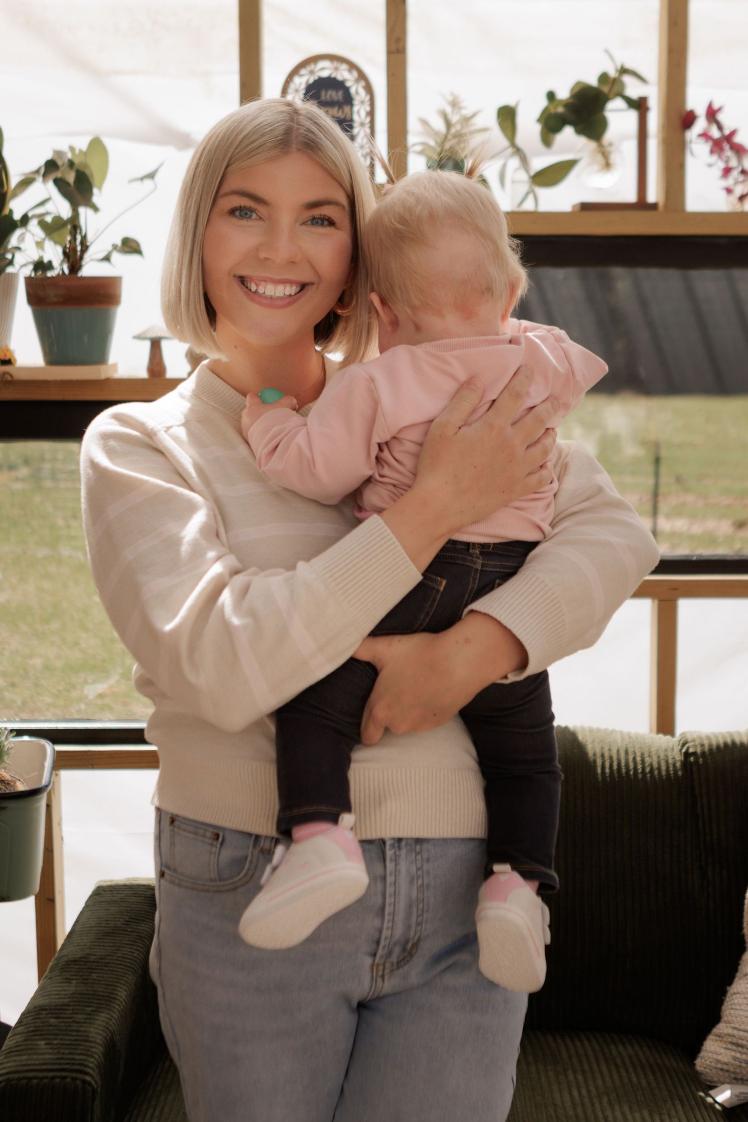 Smiling blonde woman holding a baby dressed in pink in a bright, plant-filled room.