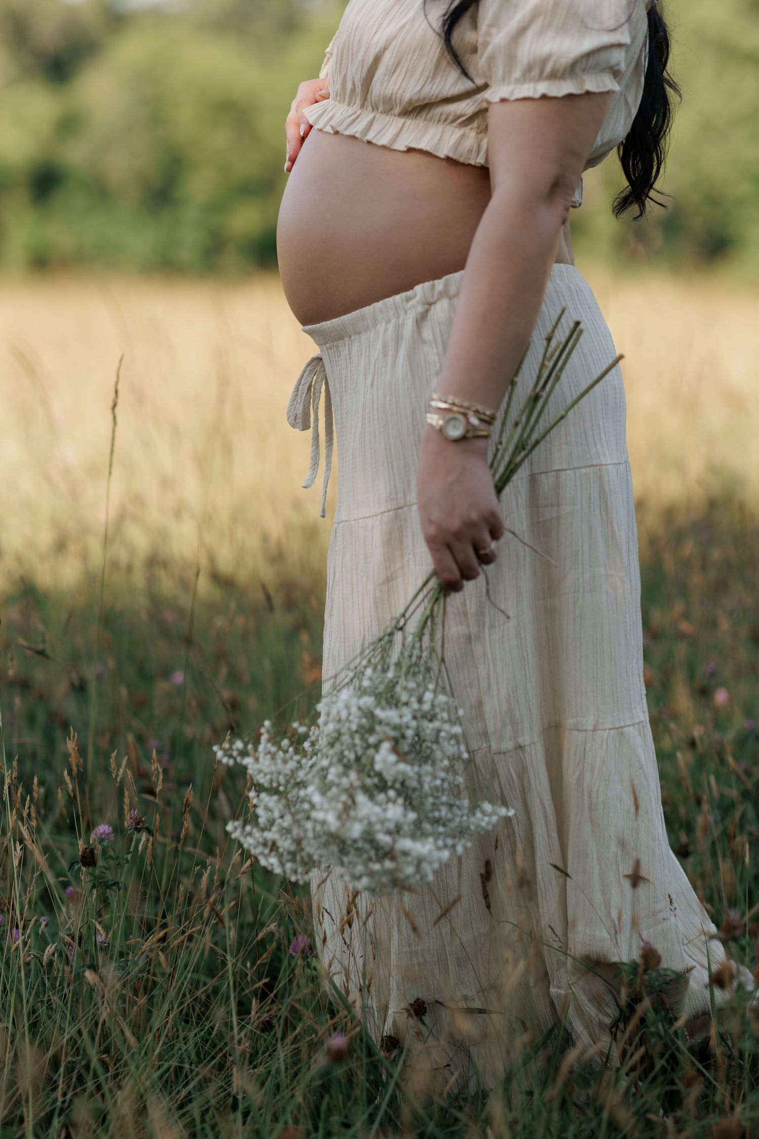 Pregnant woman in a cream outfit with exposed baby bump holds a bouquet of white baby's breath flowers in a sunlit golden field.