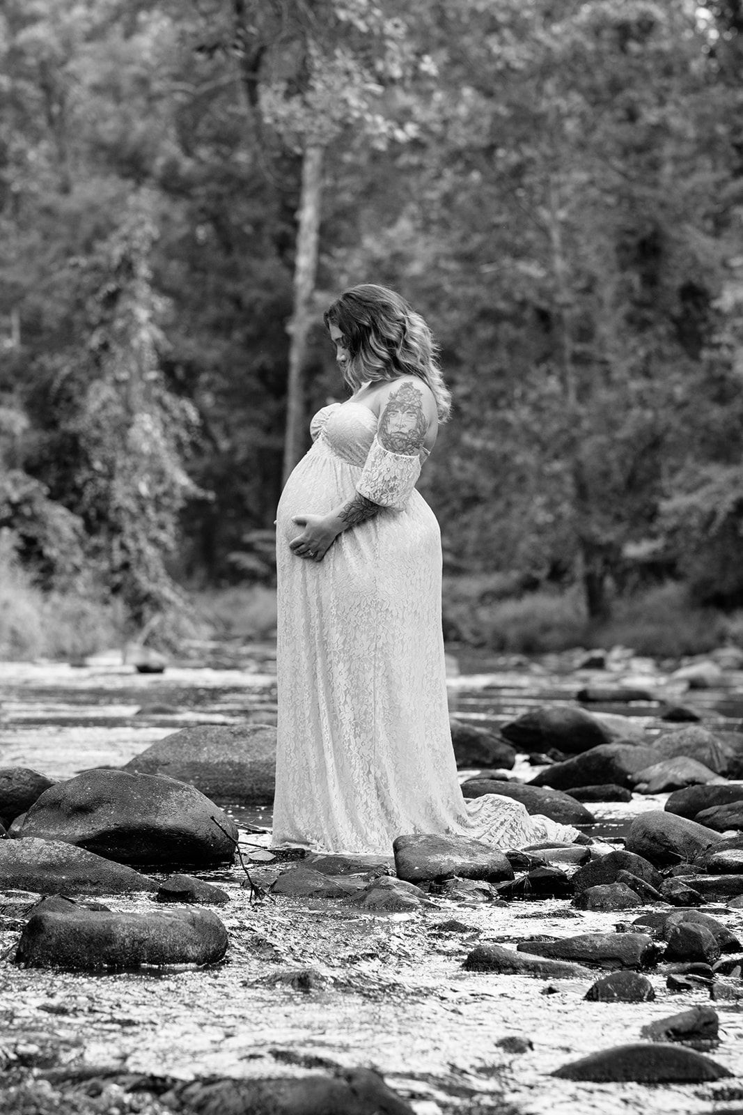 Black and white maternity portrait of a pregnant woman in a flowing lace gown standing on rocks in a shallow forest stream.