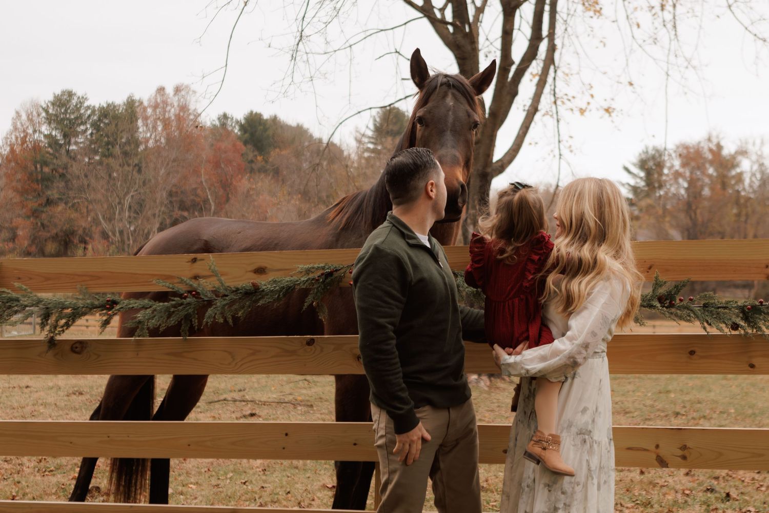 A curious horse peeks over a wooden fence at a couple in an autumn farm setting.