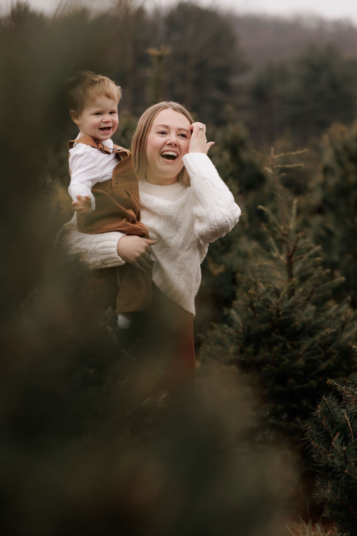 A laughing mother holds her toddler child at a Christmas tree farm, both wearing cozy winter outfits.