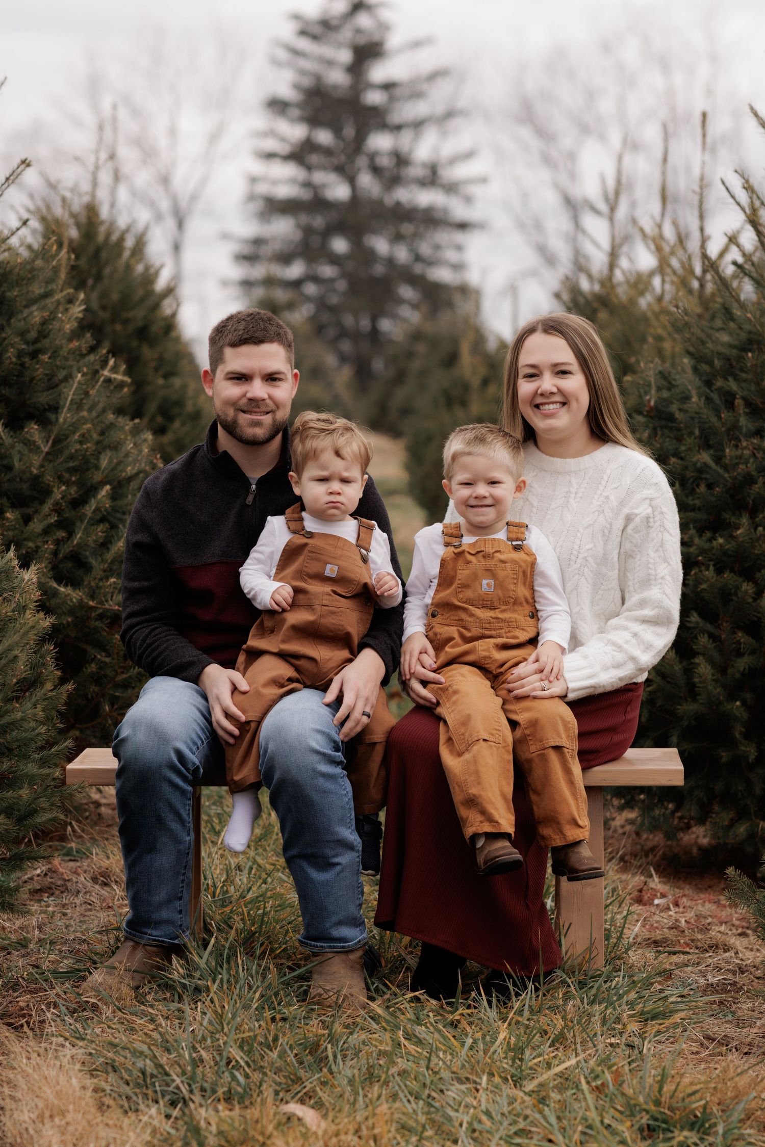 Family sitting together on rustic bench at Christmas tree farm wearing coordinated autumn outfits.