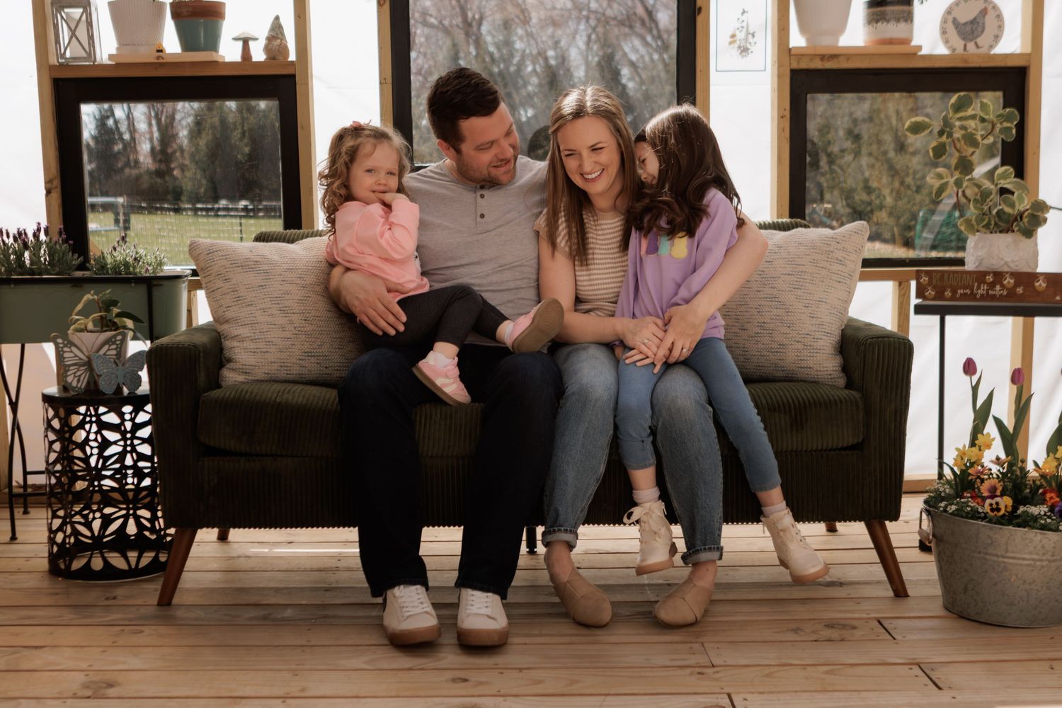 Happy family of four sitting together on a dark sofa in a cozy decorated living room with wood floors.