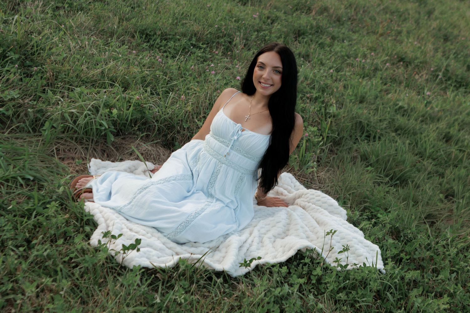 Young woman with long dark hair sitting in green grass field wearing a light blue strapless maxi dress, smiling.