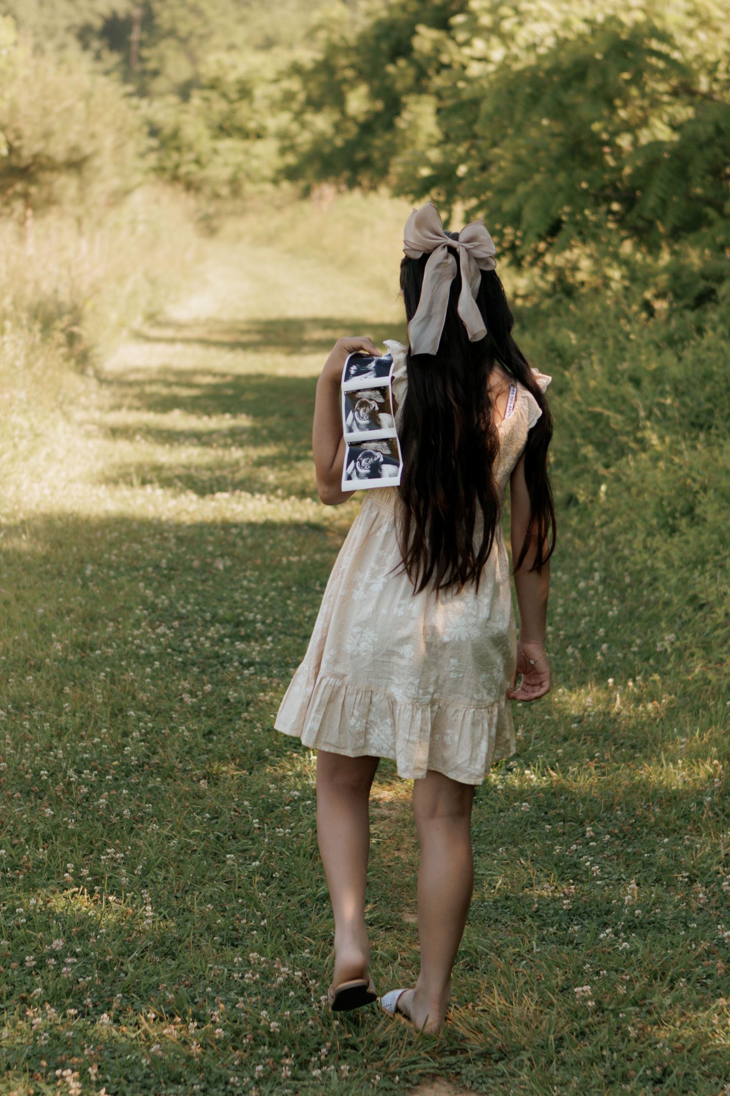 A woman in a floral dress holds ultrasound photos on a sunlit garden path, announcing her pregnancy.