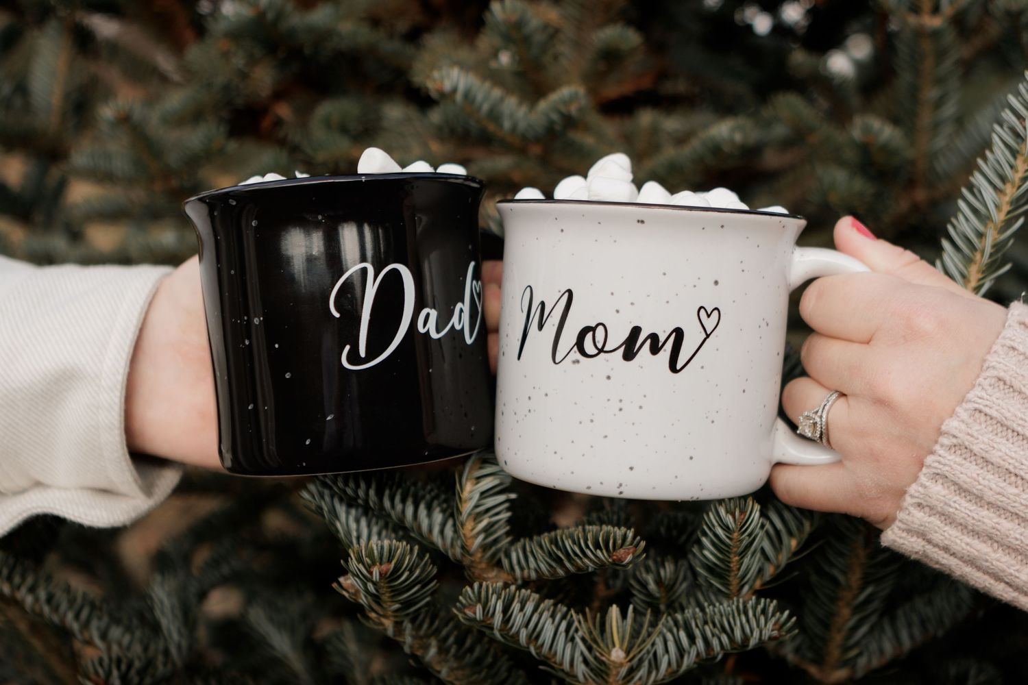 Black and white enamel mugs with 'Dad' and 'Mom' text displayed among pine branches.