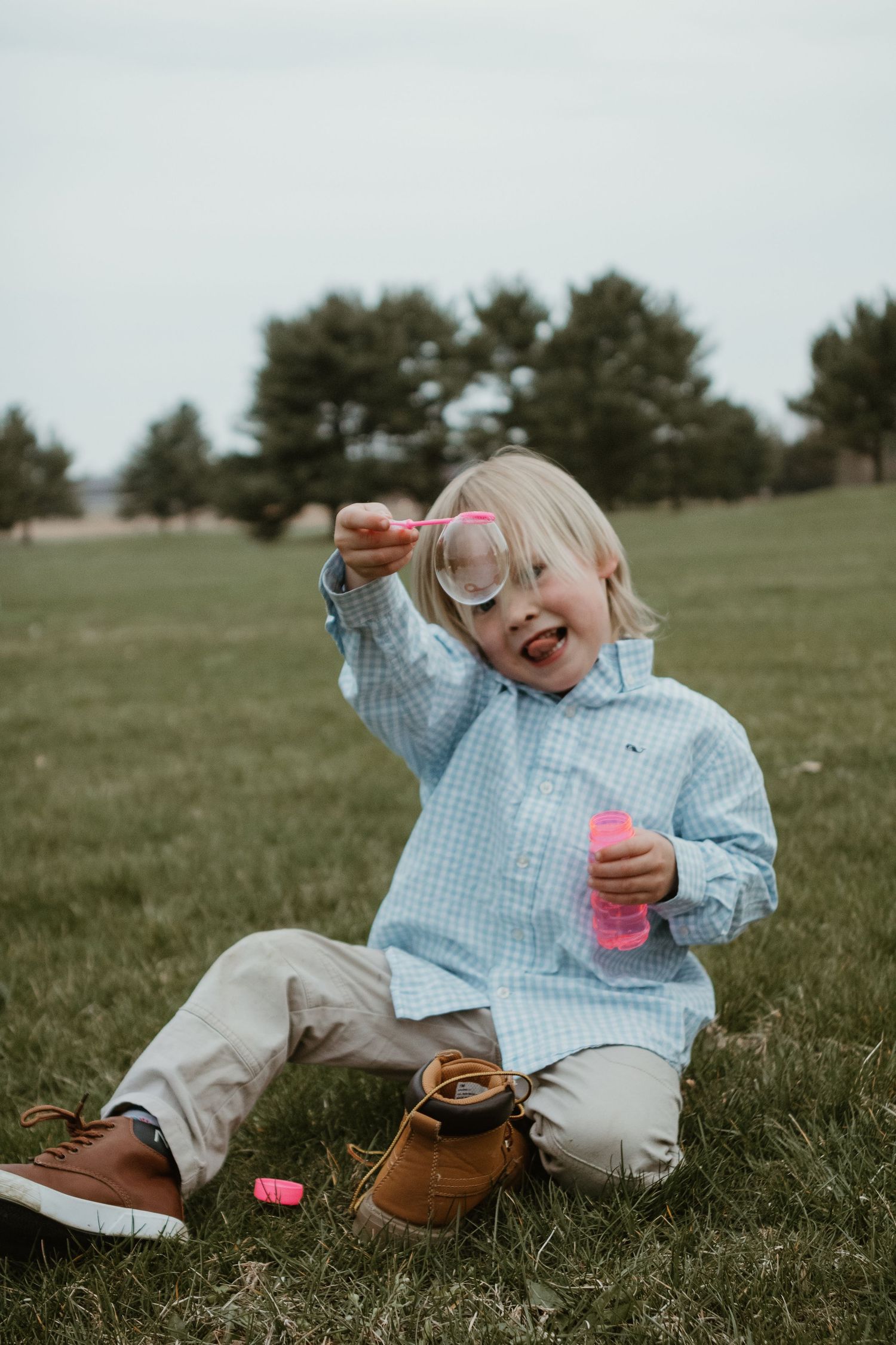 A small child in a blue gingham shirt and khaki pants sits playfully on grass while laughing and pointing.