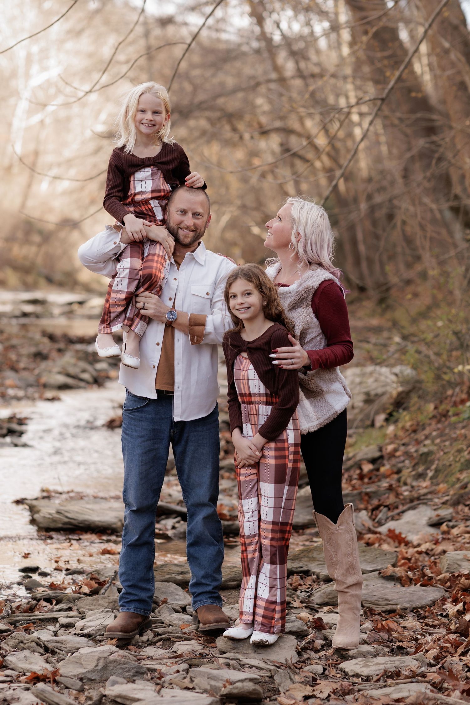 Family in coordinated plaid and burgundy outfits posing on stone path surrounded by fall leaves.
