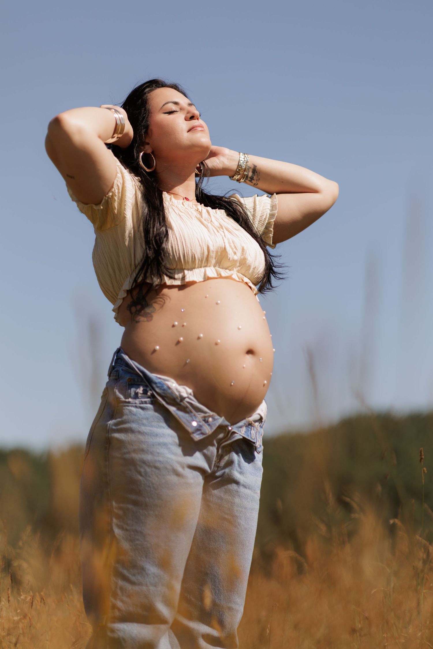 Pregnant woman with long dark hair and arms raised stands in a golden field under a blue sky, her bare belly decorated with small pearls.