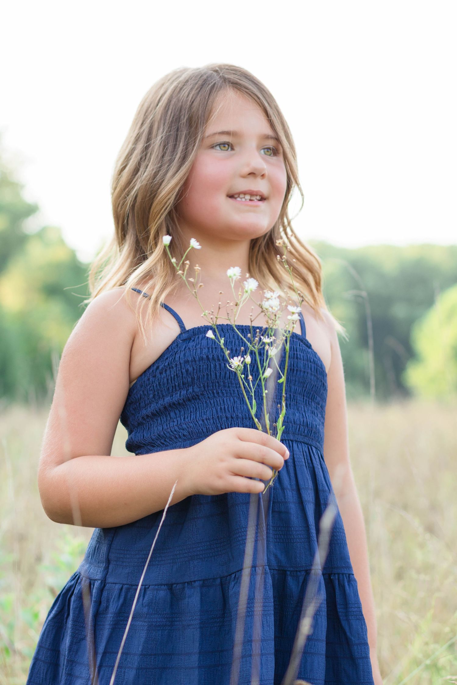 A young person in a navy blue sundress holds dried wildflowers while standing in a sunlit meadow.