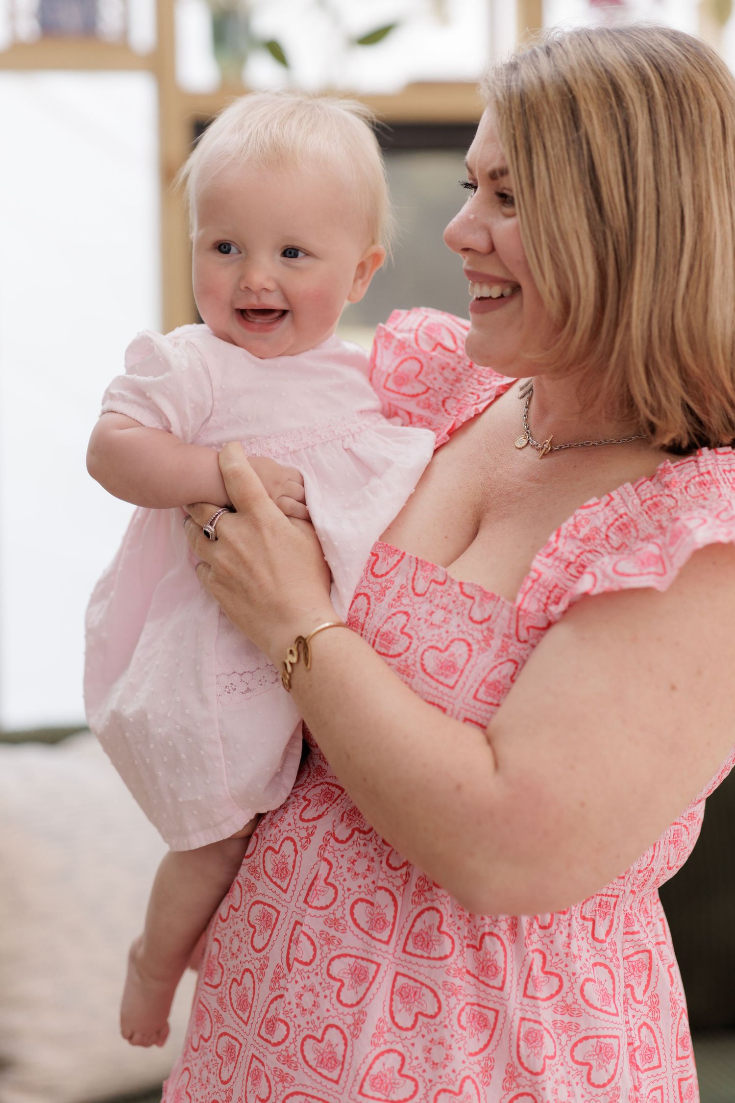 A smiling woman in a pink heart-print dress holds a happy baby girl in a light pink dress.