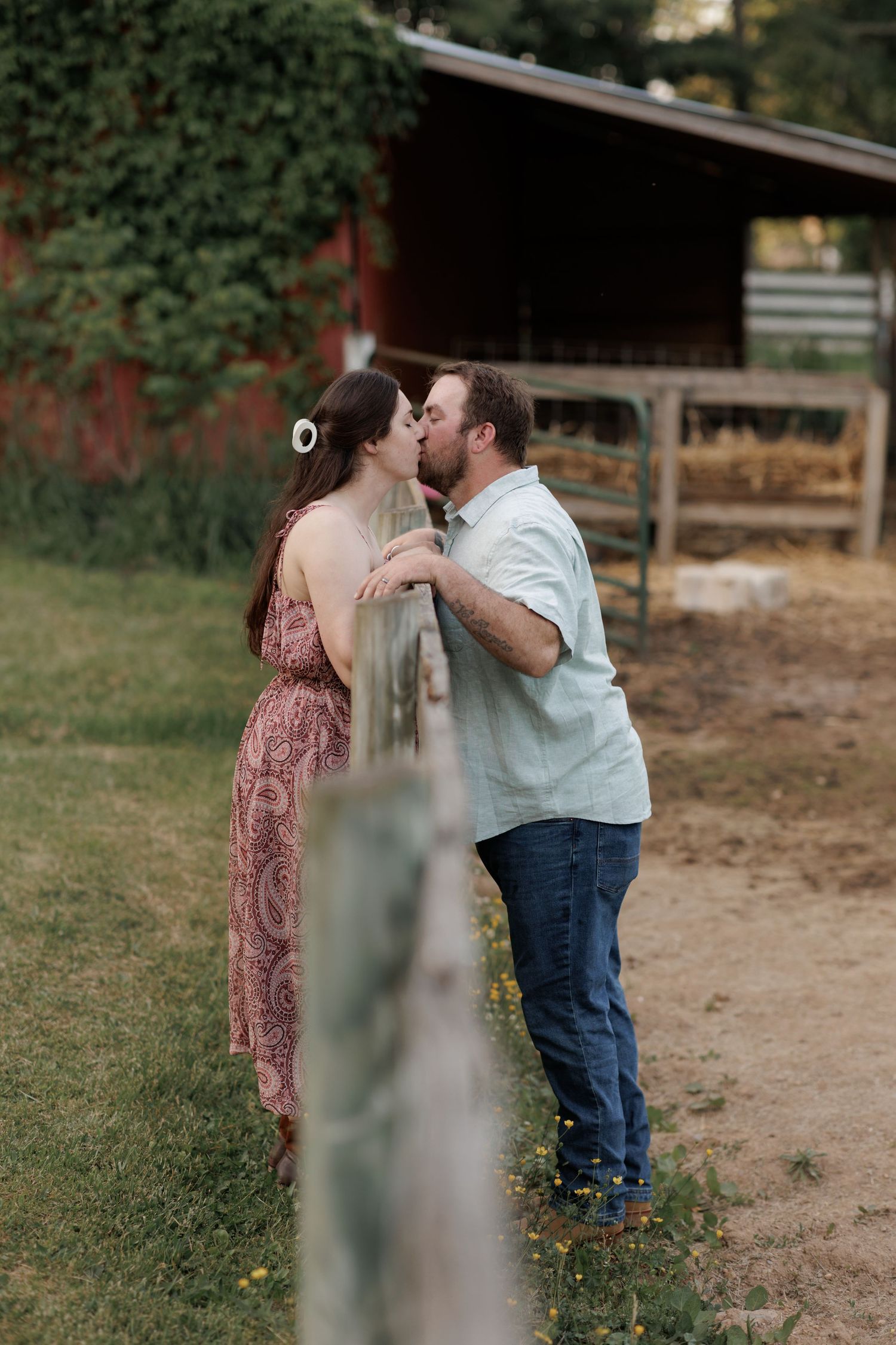 Couple sharing a romantic kiss by a rustic wooden fence on a farm, woman in floral dress, man in light blue shirt.