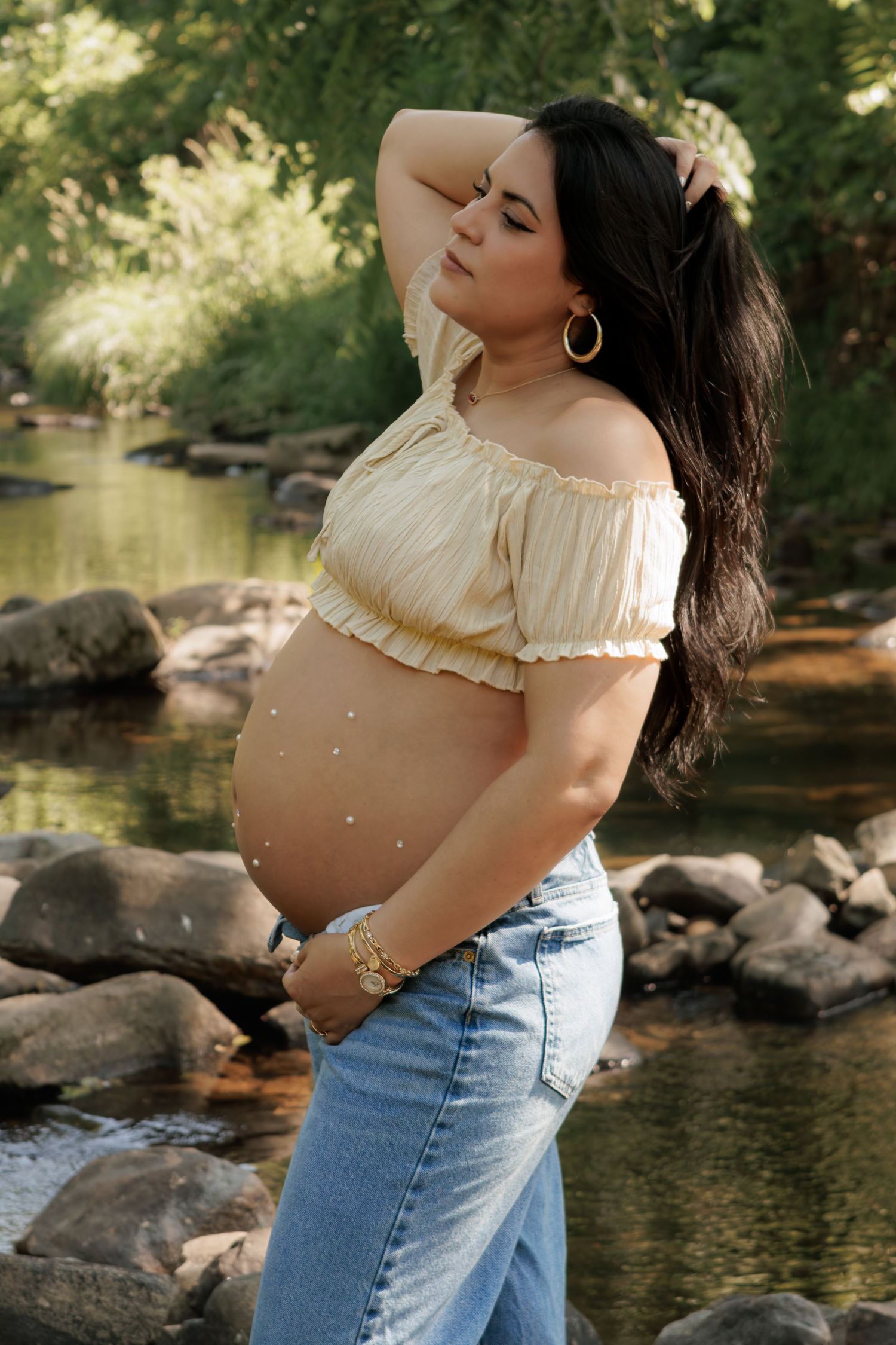 Pregnant woman in a cream off-shoulder top and denim jeans poses with her hand in her hair beside a rocky, sunlit stream.