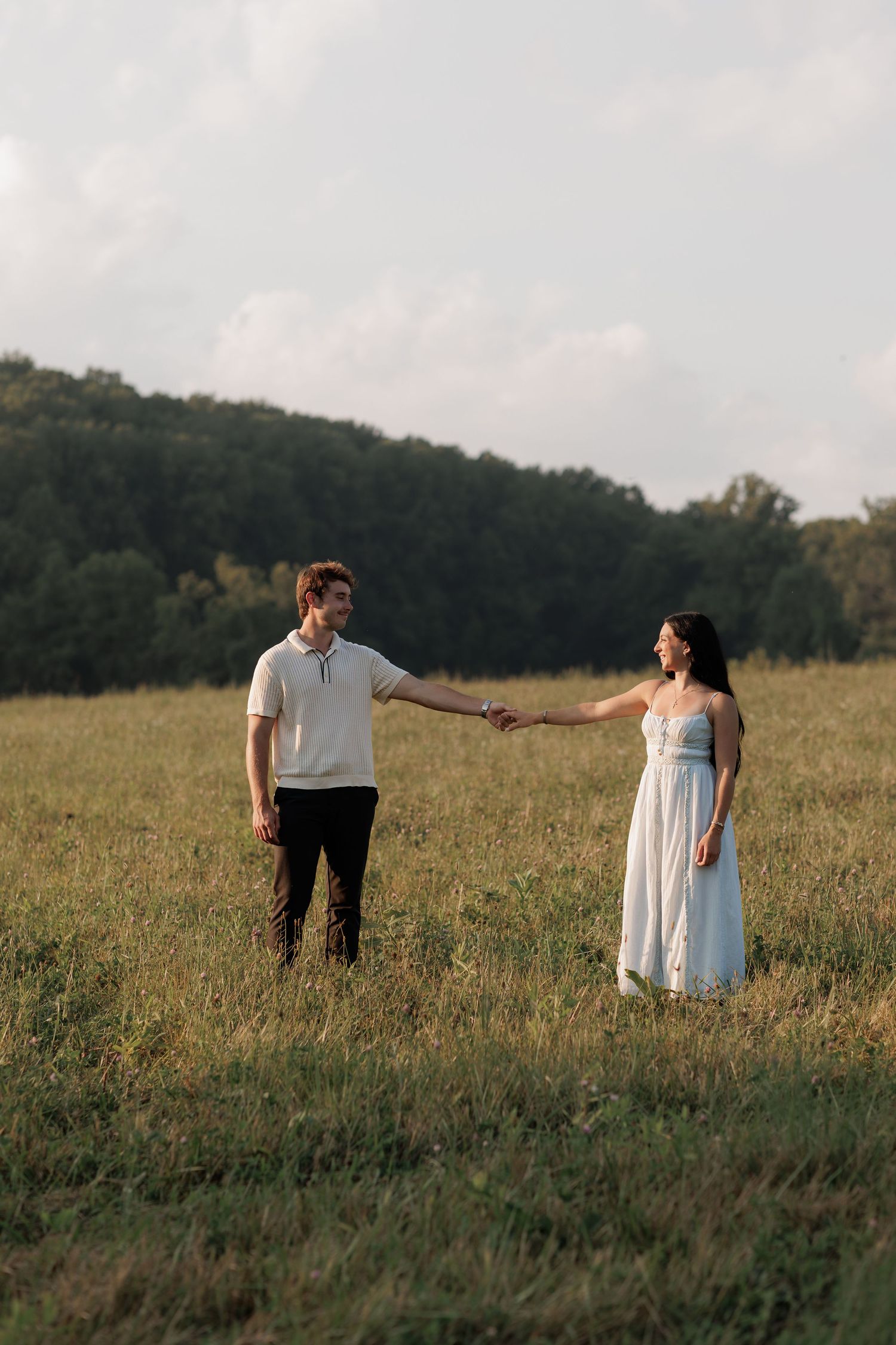 A couple holds hands in a sunlit green field with trees in the background, the woman in a white dress.