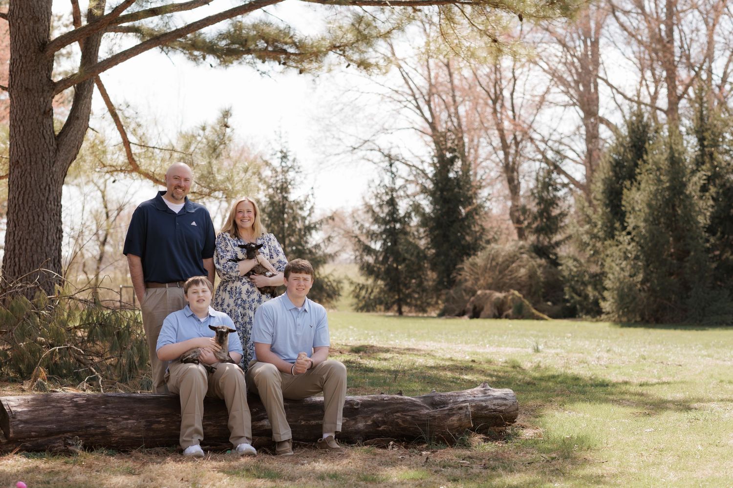 Family of four posing outdoors on a log in a lush green park surrounded by tall trees.