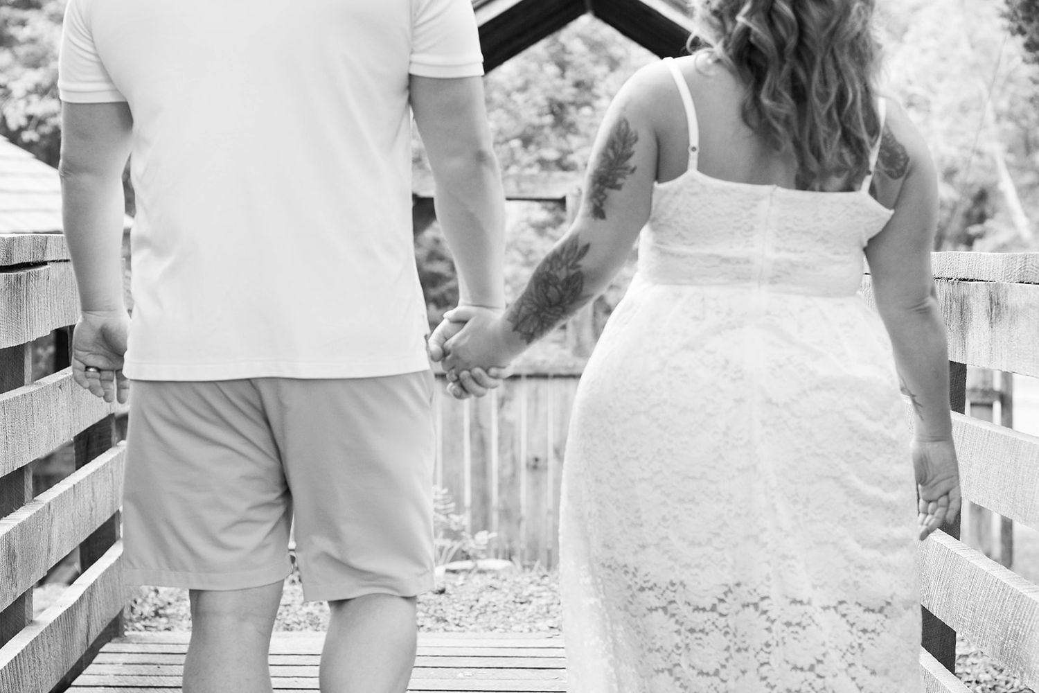 Black and white photo of a couple holding hands on a wooden bridge, woman in lace dress with tattoo.