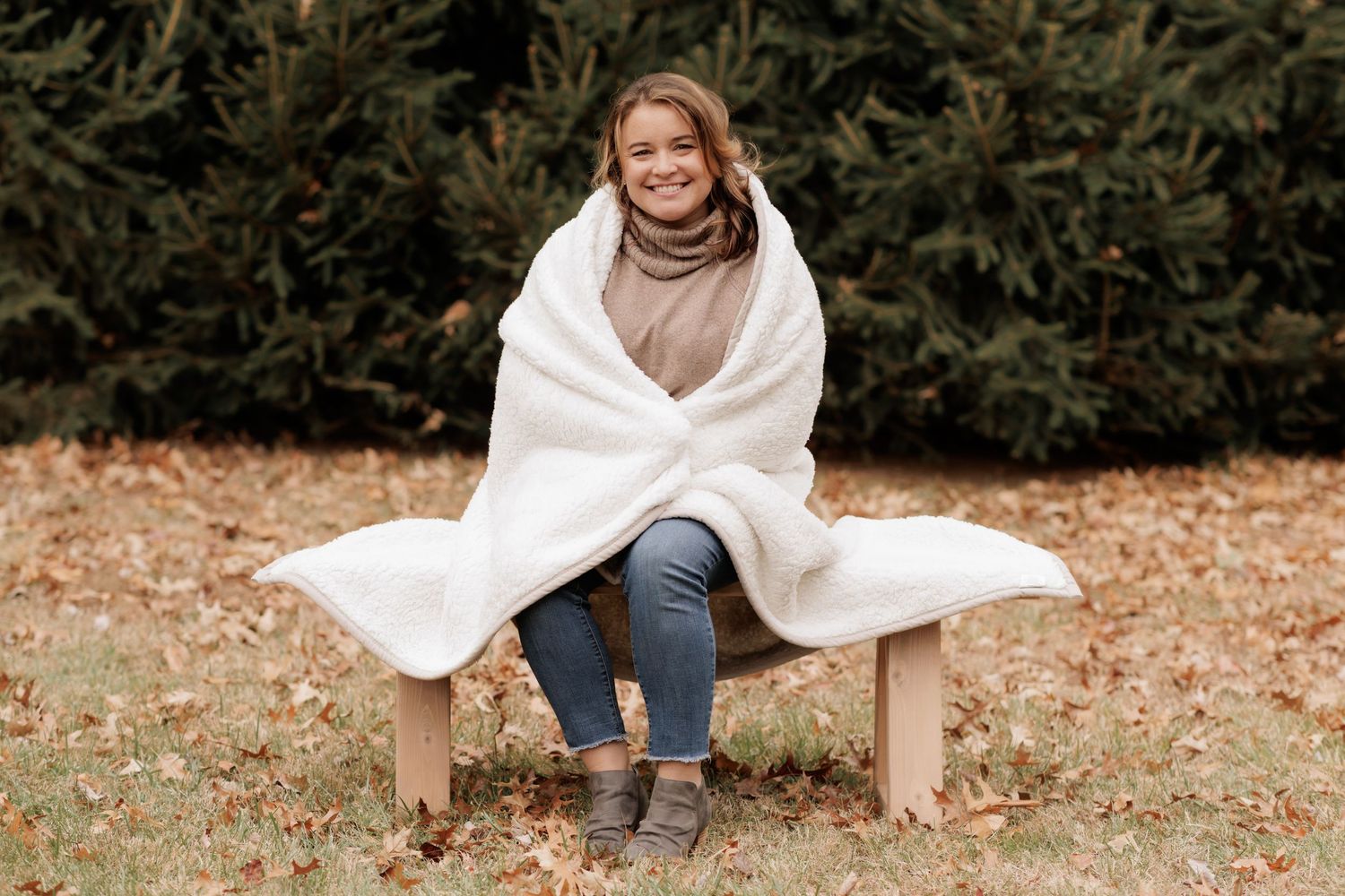 Person wrapped in white blanket sits smiling on wooden bench near evergreens.