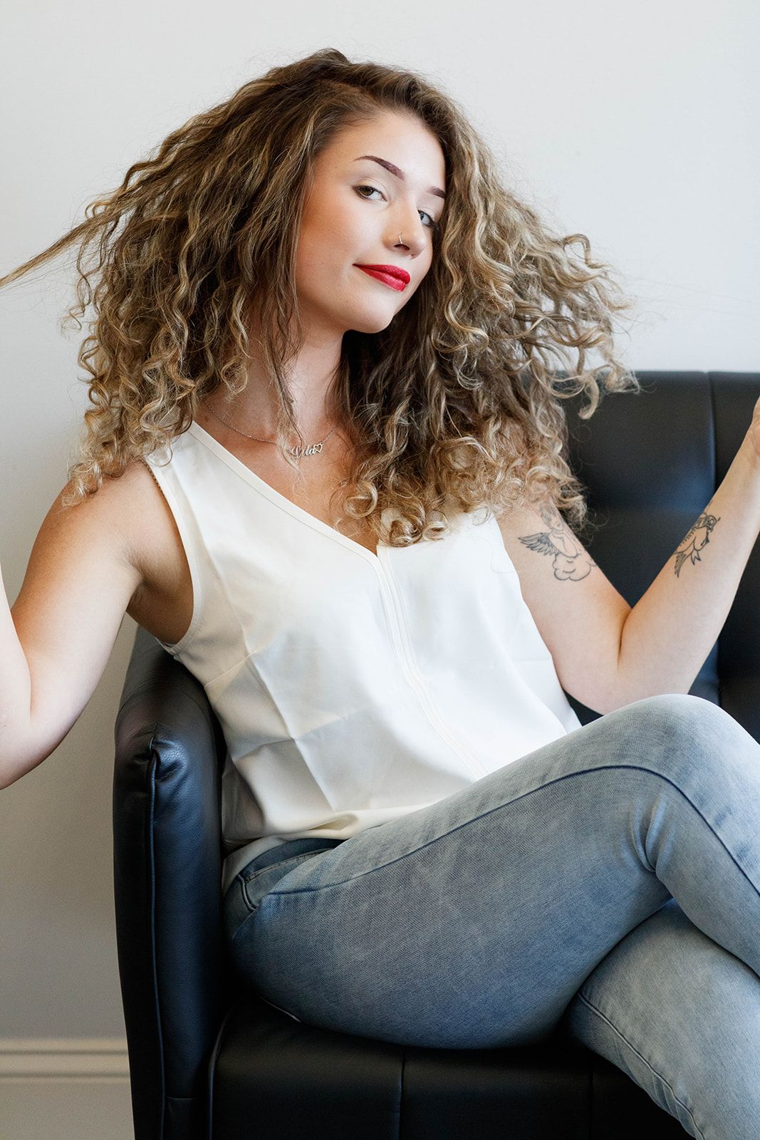 A person with curly hair in white tank top poses confidently on black leather chair.