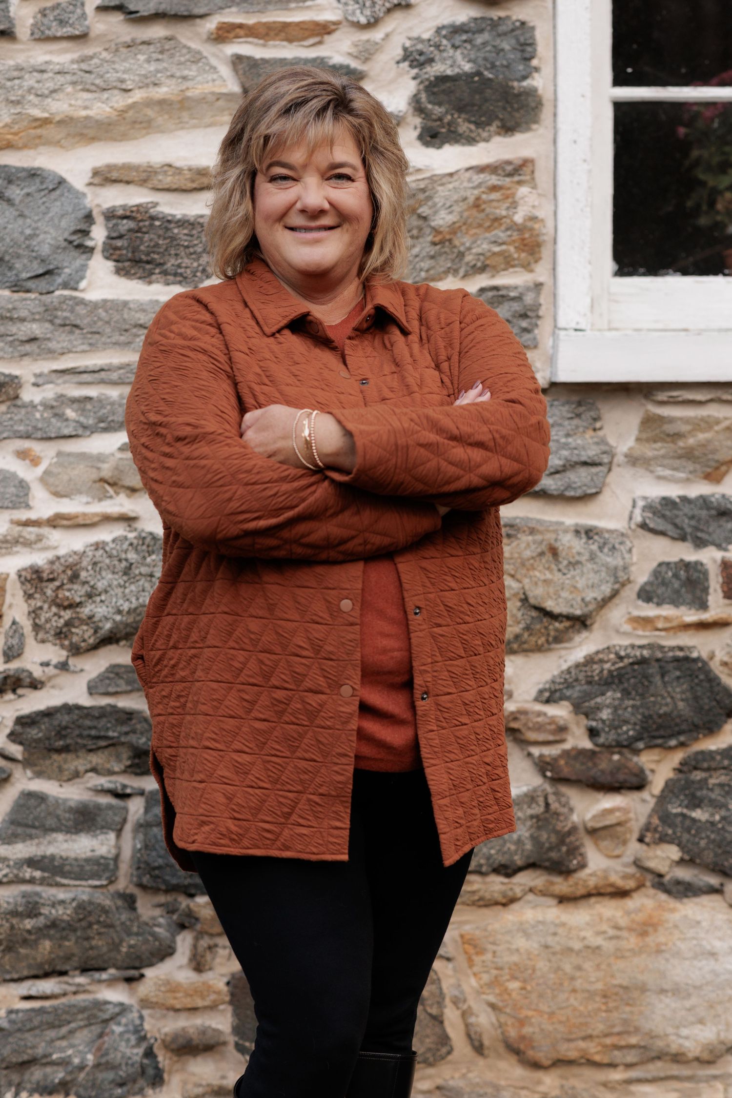 Woman in rust colored quilted jacket smiles against stone wall.