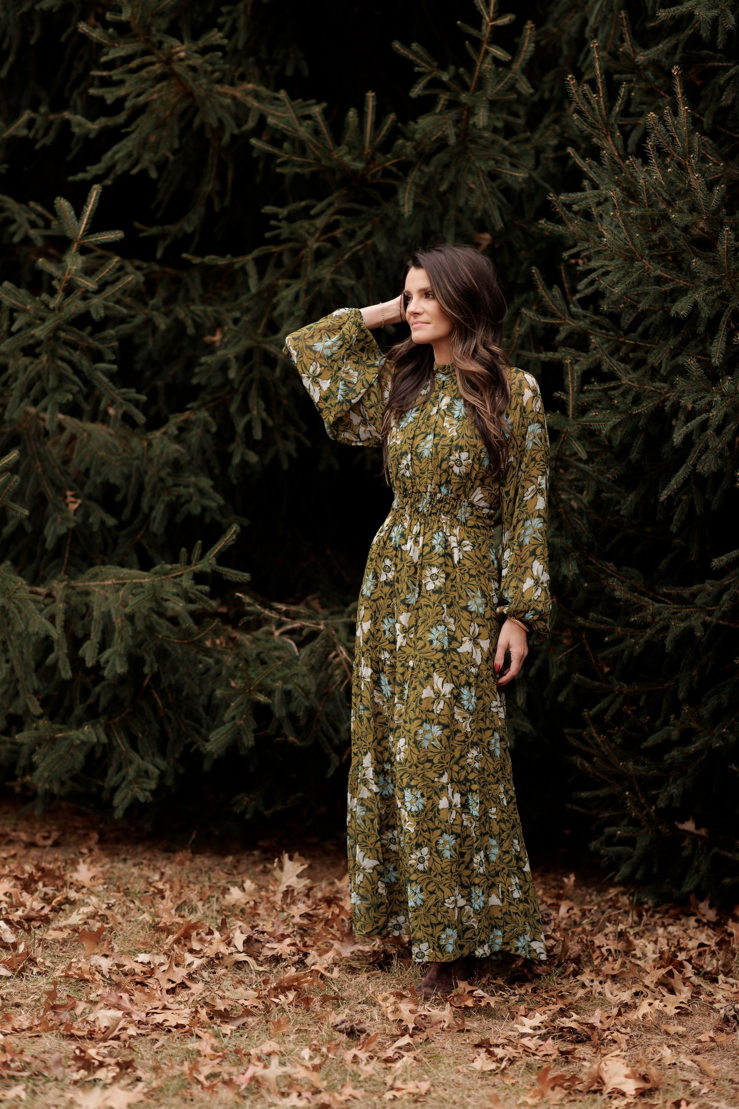 Woman in long floral maxi dress poses against evergreen trees in autumn.