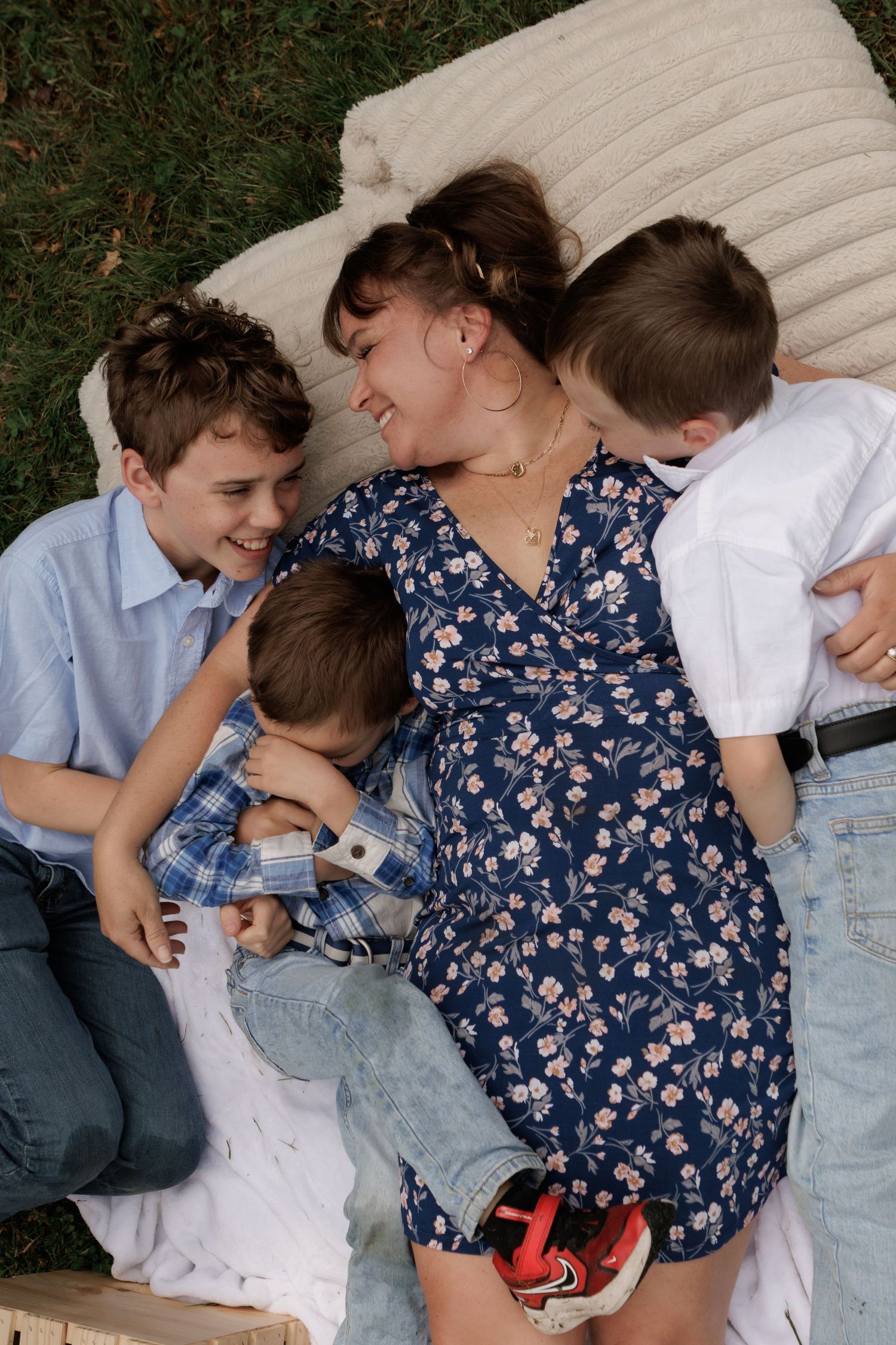 A mother laughing with her three young sons while relaxing together on a blanket in the grass outdoors.