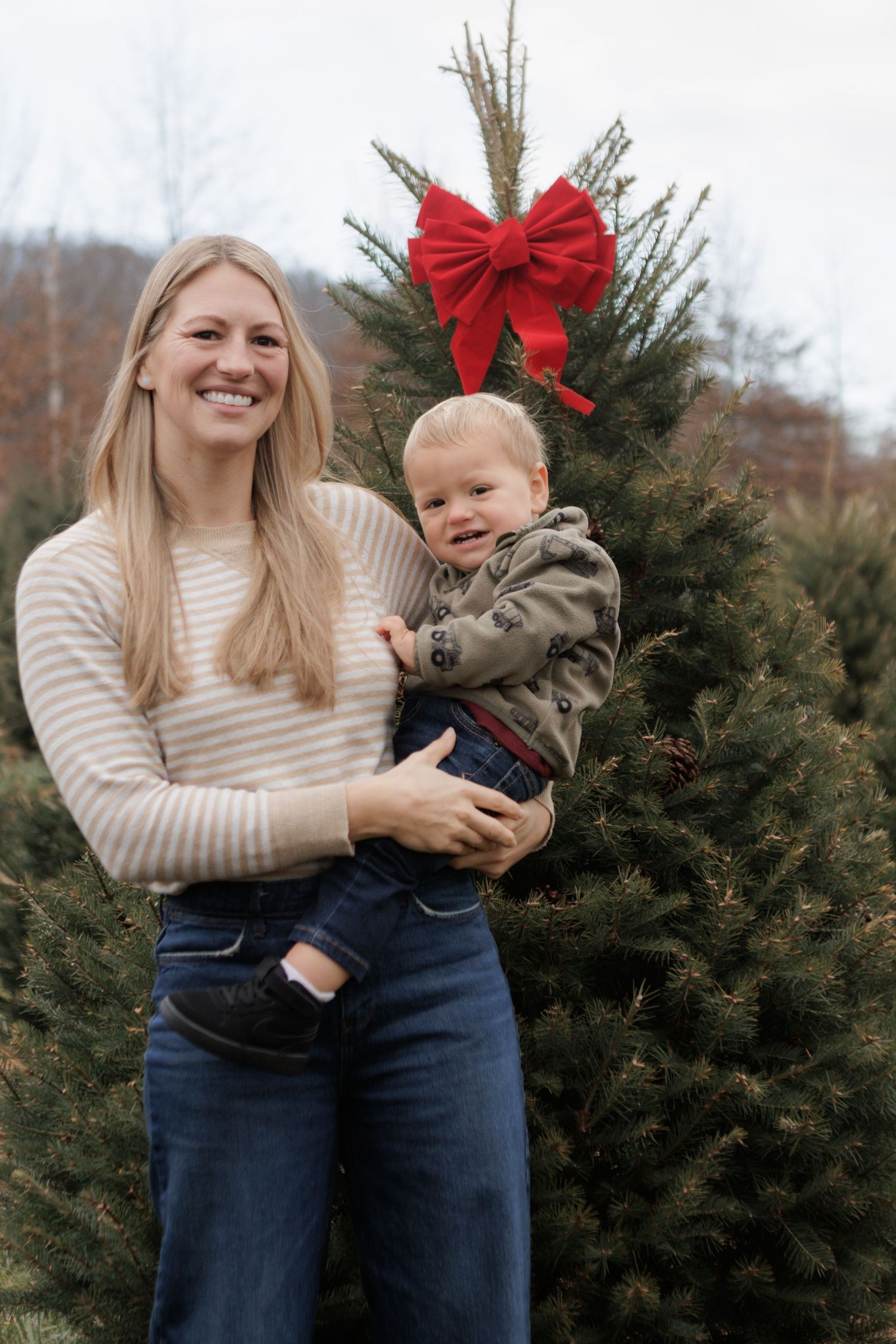 A smiling woman holds a toddler boy in front of a Christmas tree with a red bow at a tree farm.