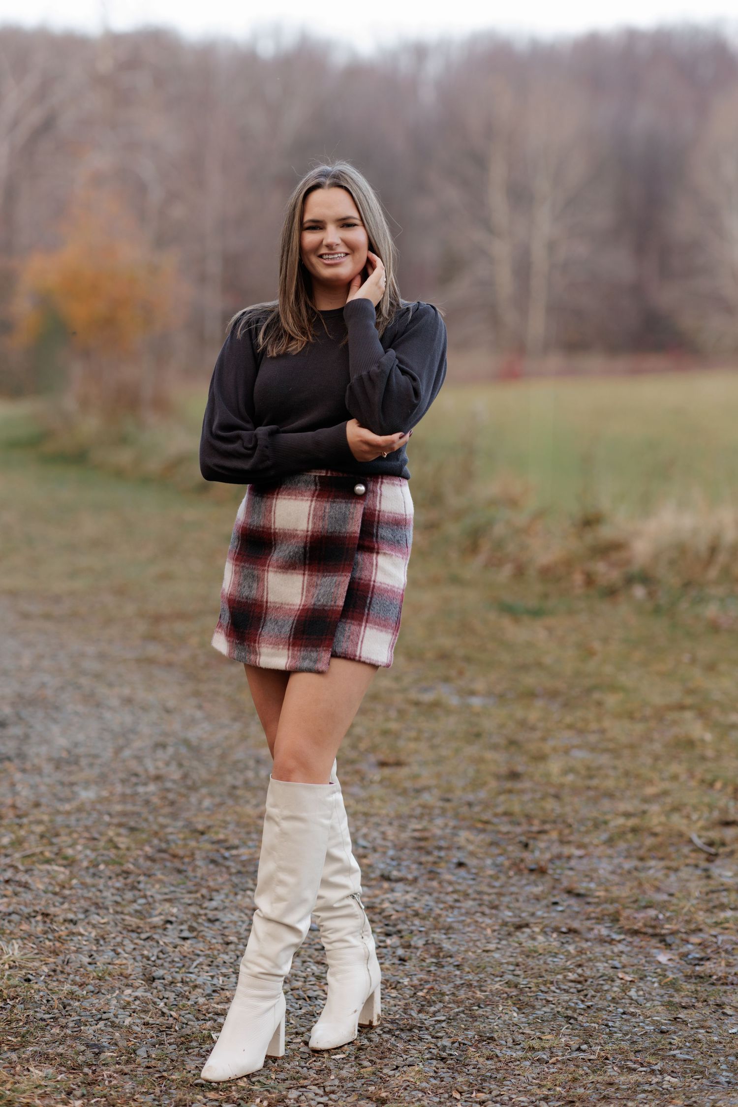 Woman in black sweater and plaid skirt with white boots poses on gravel path.