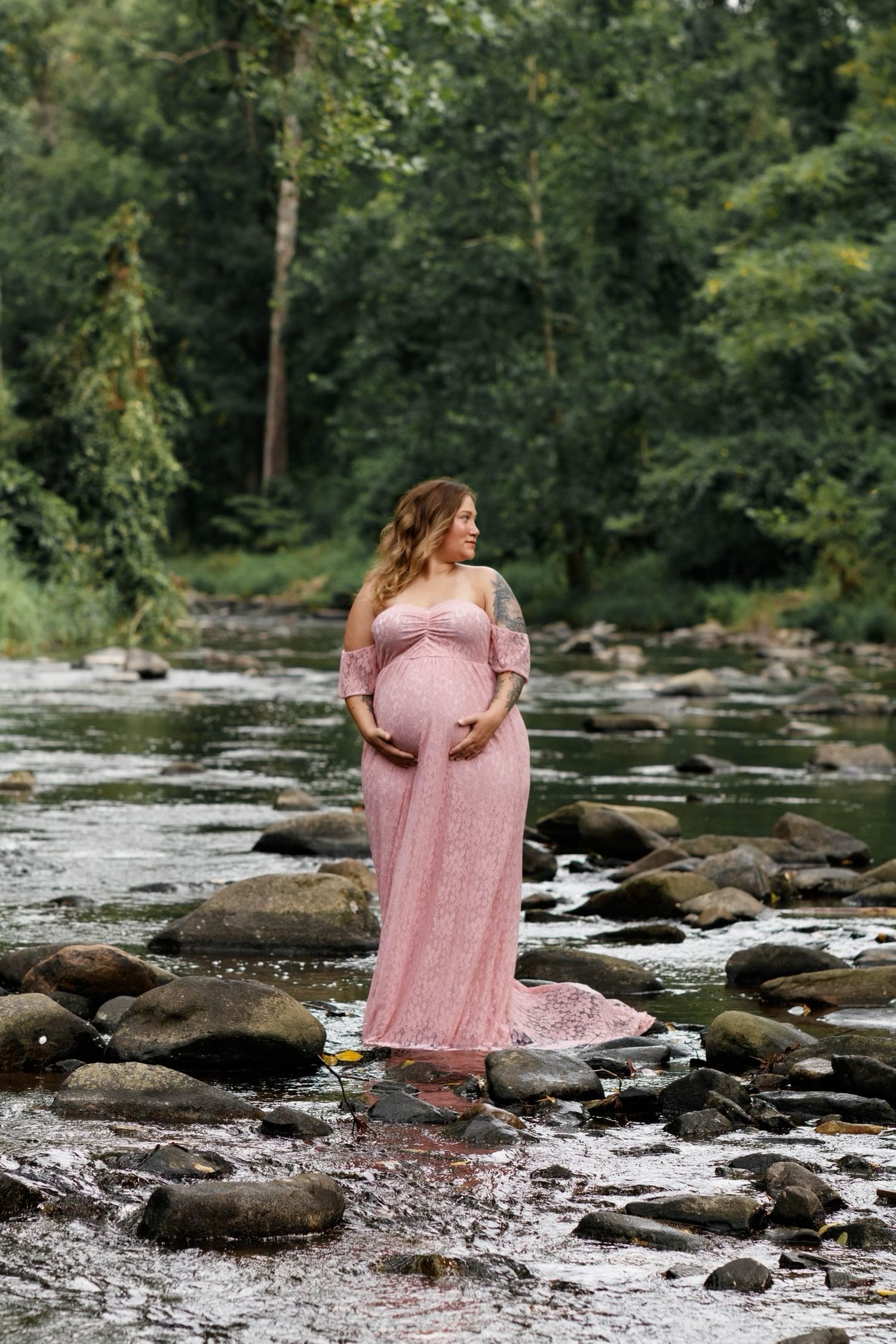 Expecting mother in pink lace dress standing on rocky riverbed surrounded by forest.