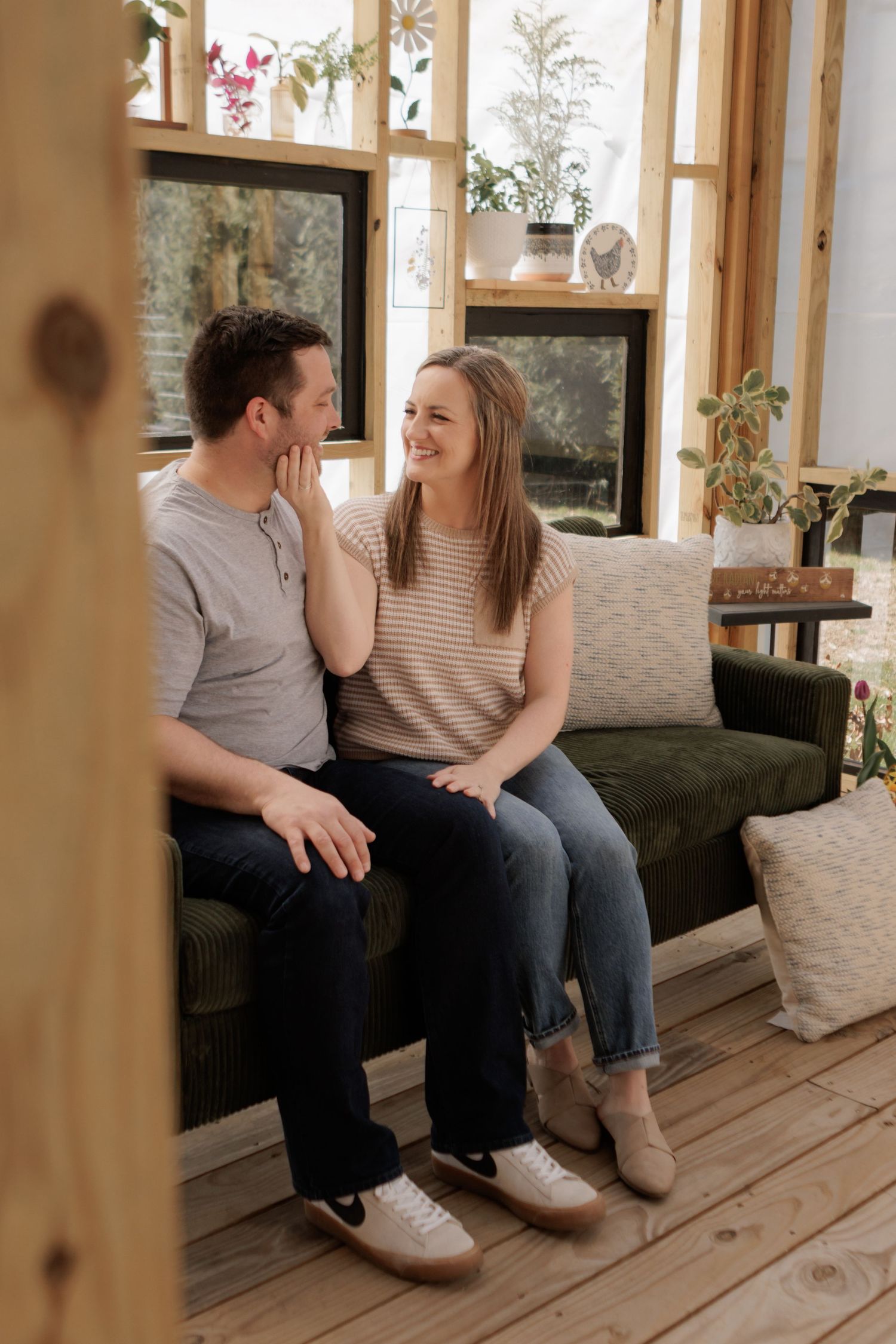 A happy couple sits together on a green sofa in a cozy sunroom, smiling and enjoying each other's company.