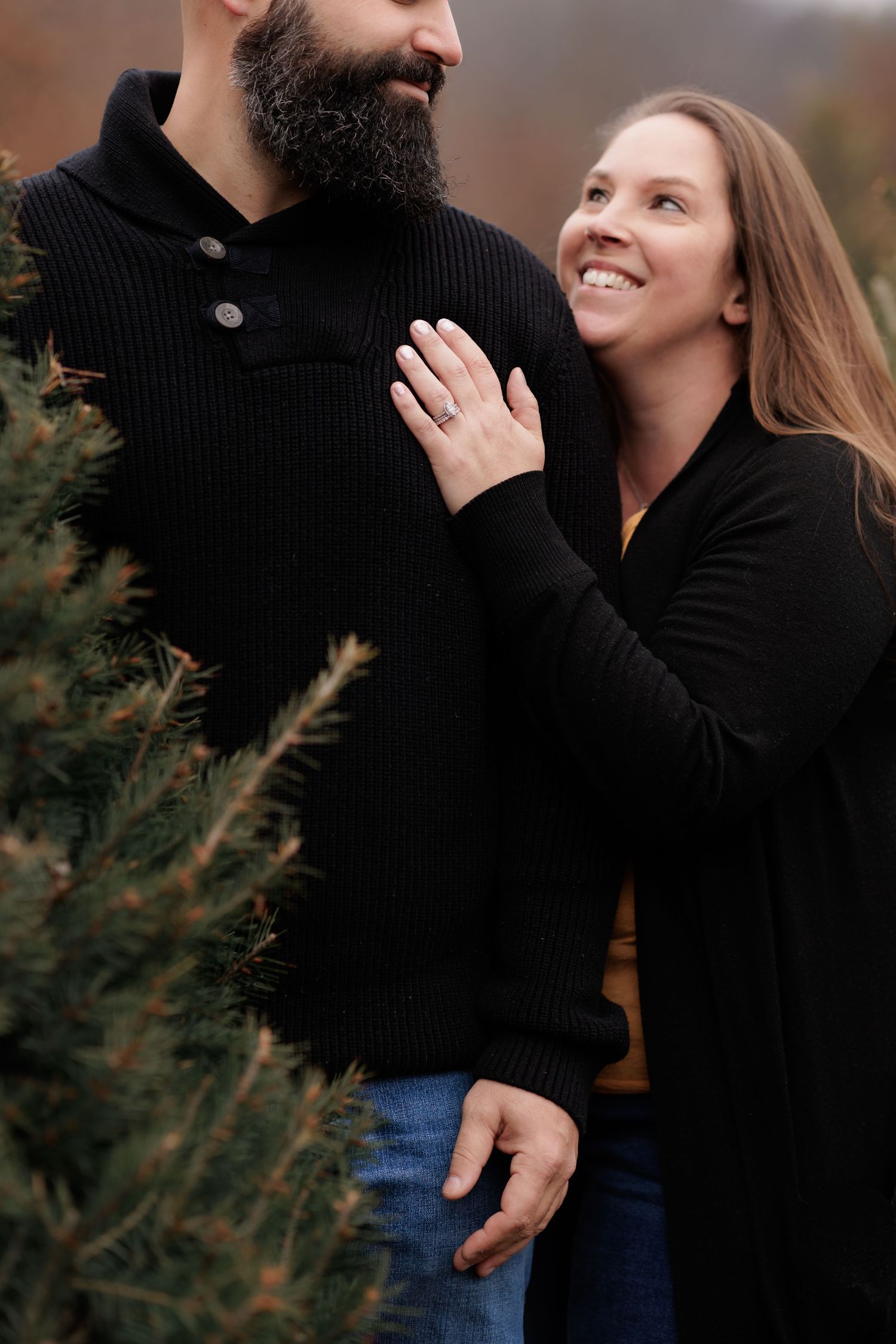 Close-up of couple showing off engagement ring among evergreen branches.