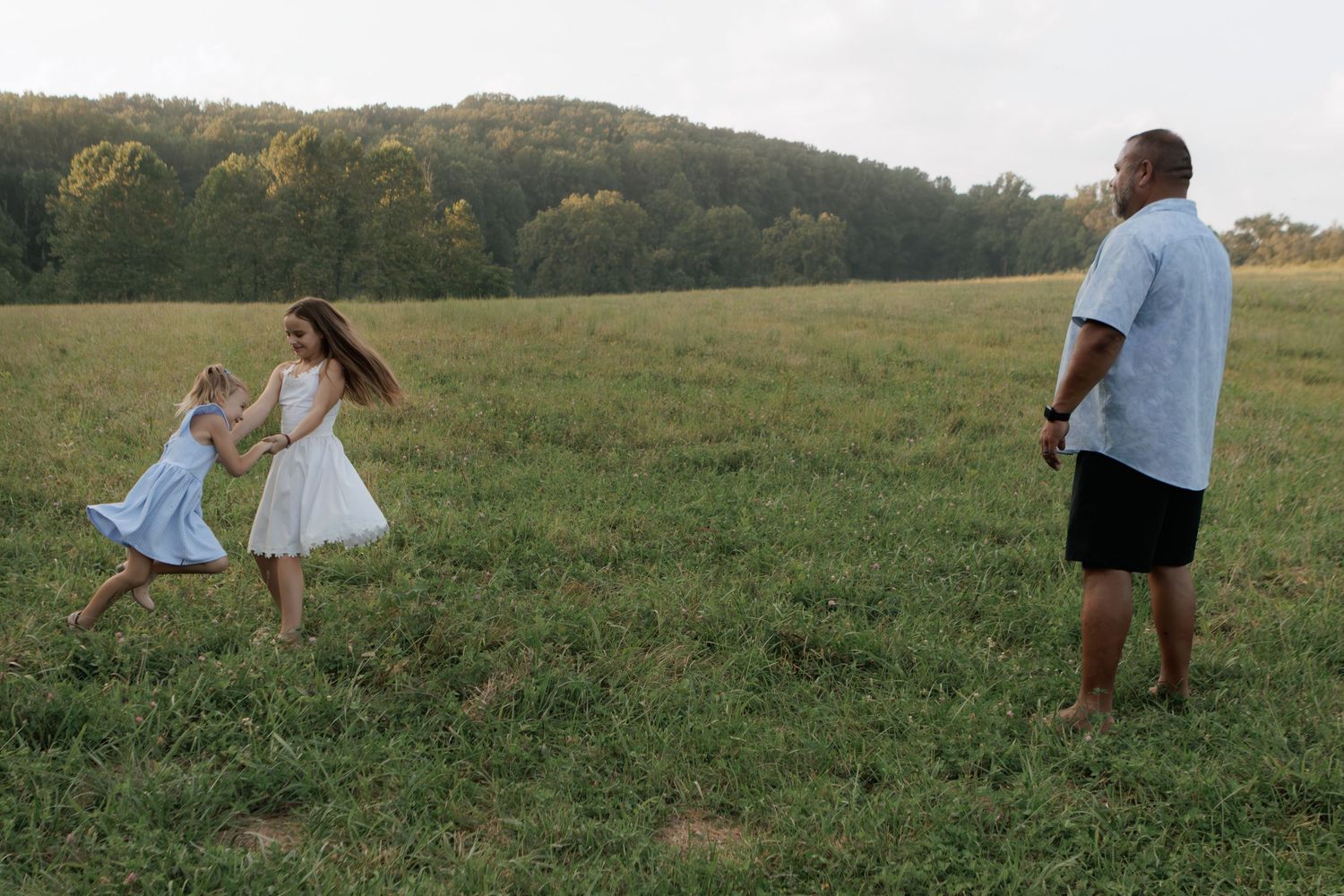 A father watches two young girls in white dresses running and playing together in a lush green open field.