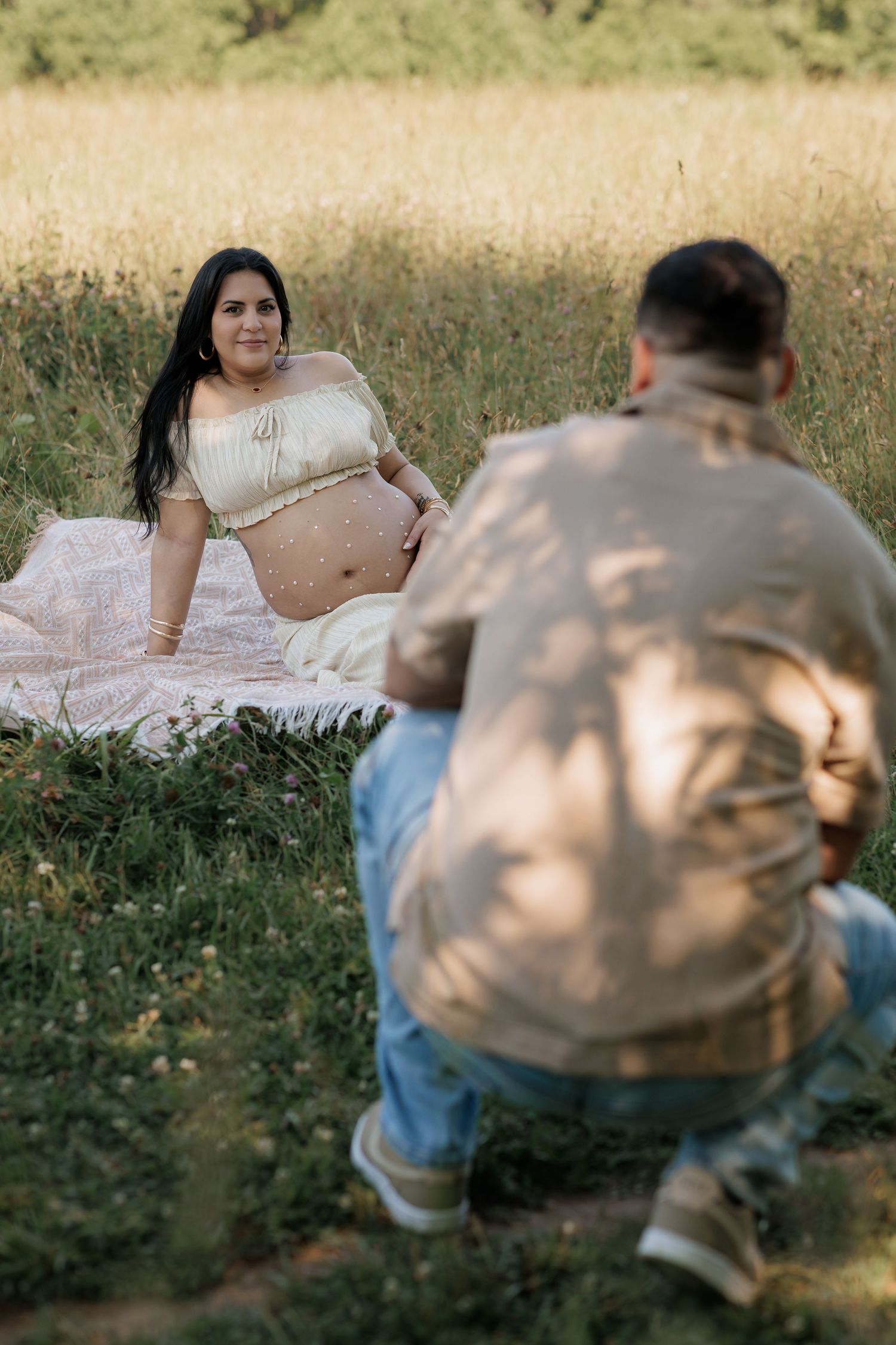 Pregnant woman sitting on blanket in field smiling at partner during outdoor maternity photoshoot.