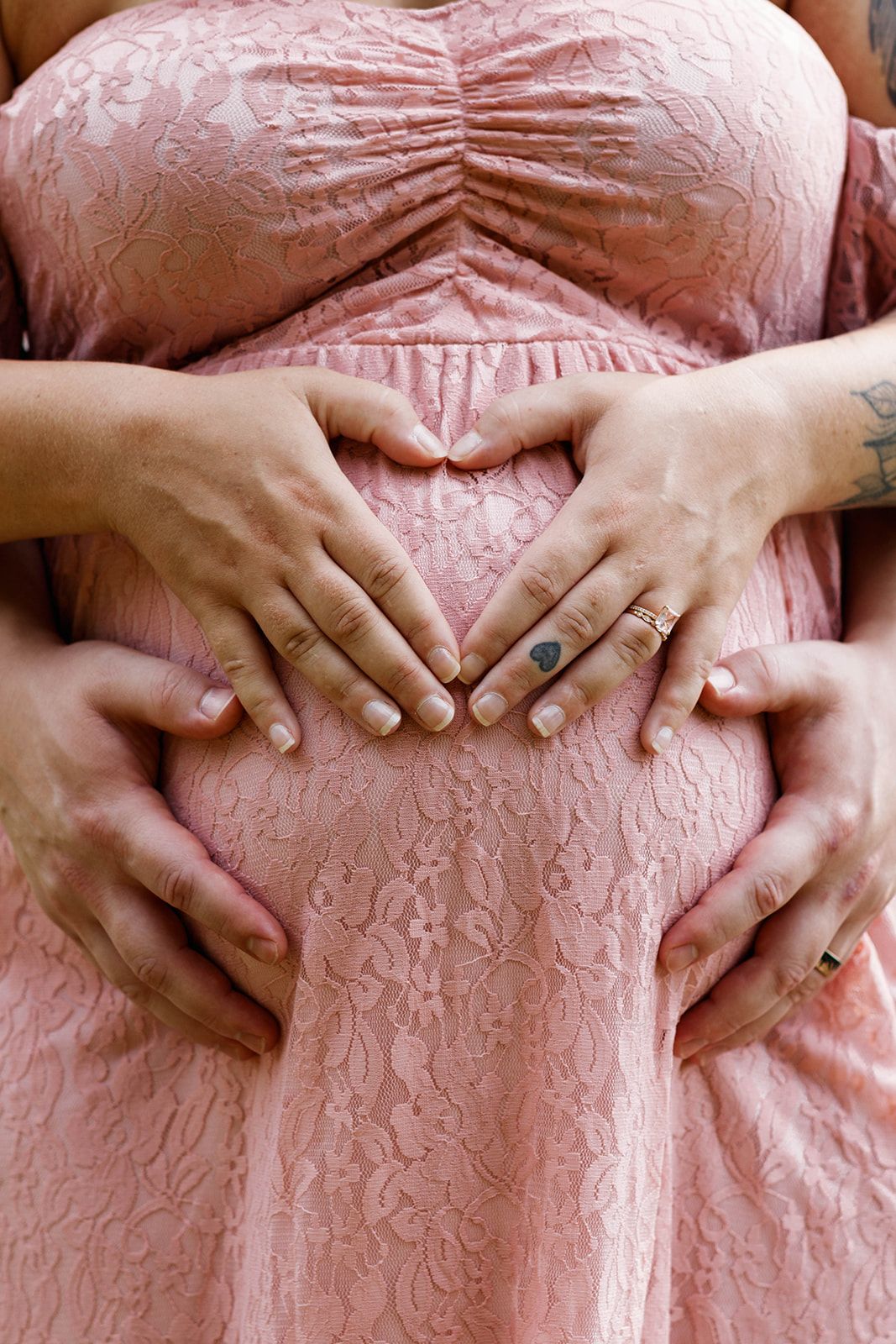 Several hands form a heart shape on the baby bump of a pregnant woman wearing a pink lace maternity dress.