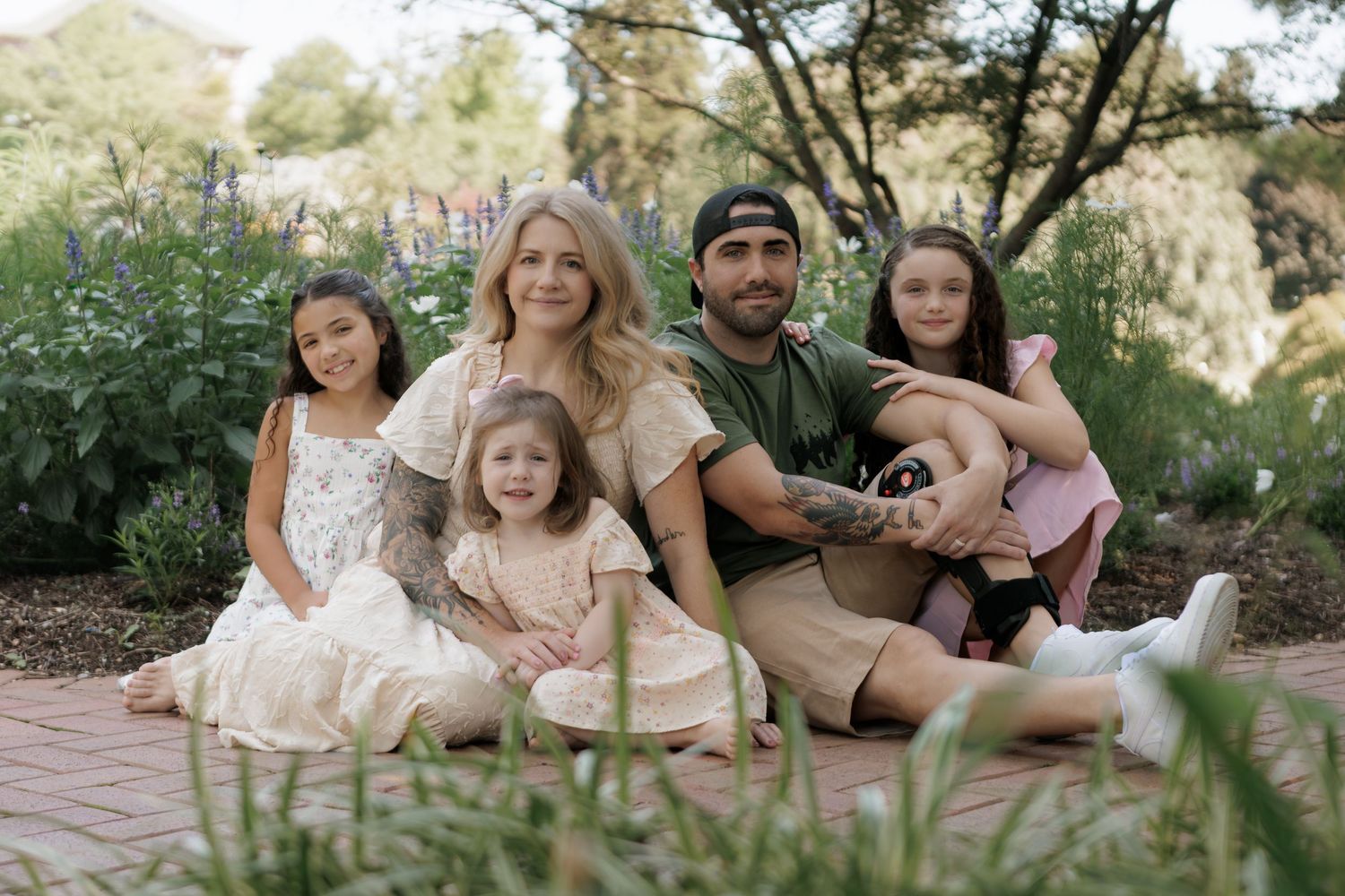 Family of five posing outdoors on brick path surrounded by lush greenery, with parents and three young daughters smiling.