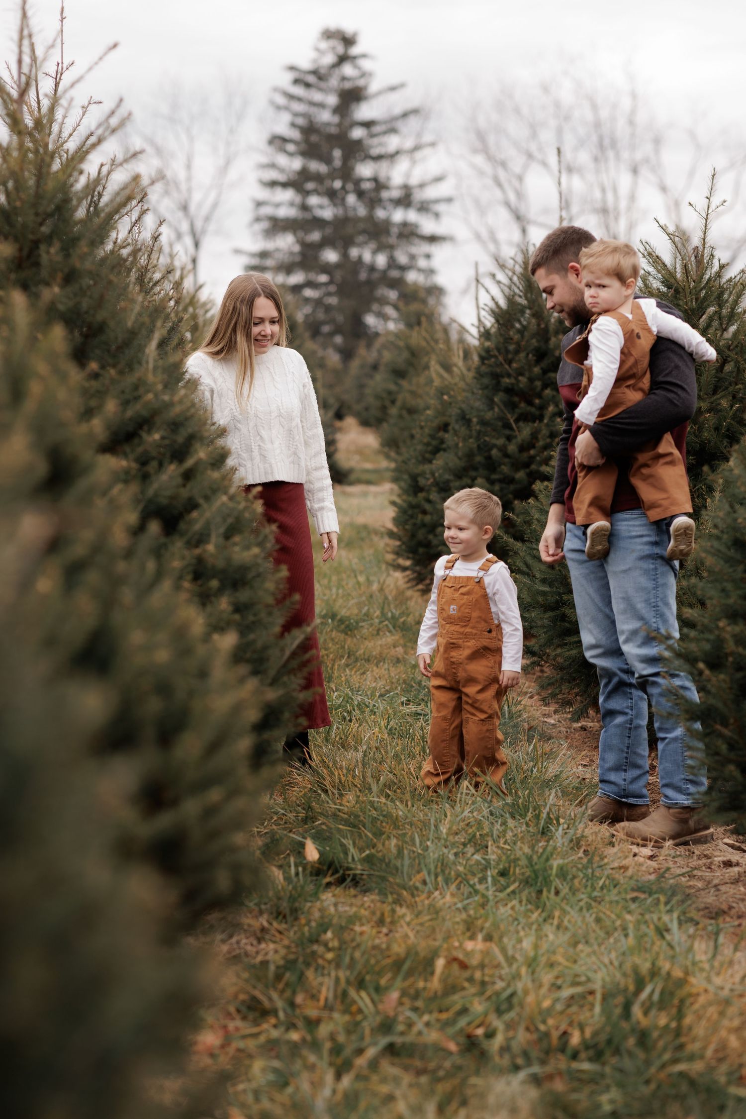 Family choosing a Christmas tree at a festive tree farm, with two young children in autumn overalls.