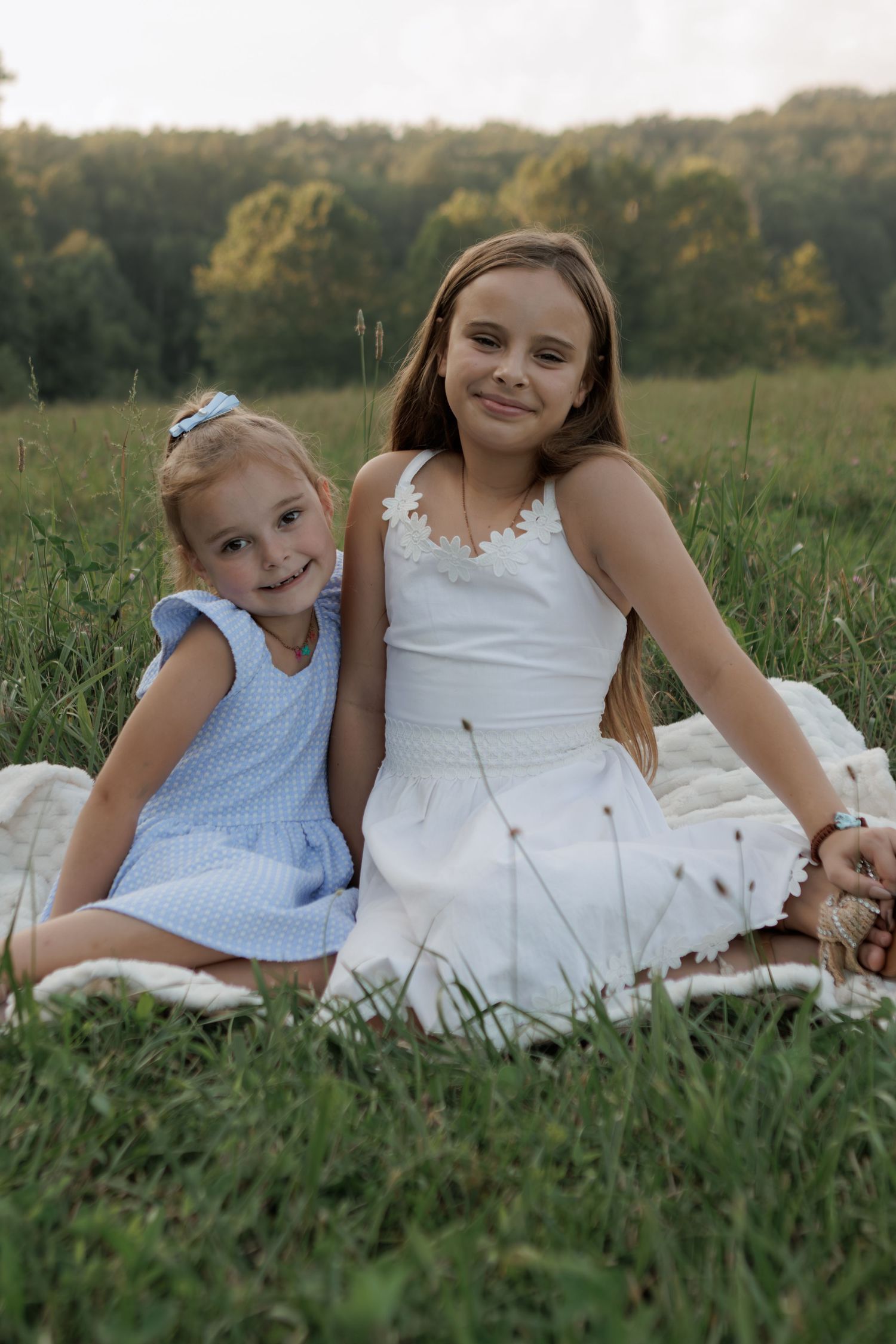 Two young girls in summer dresses sitting on a blanket in a green field, smiling warmly at the camera.