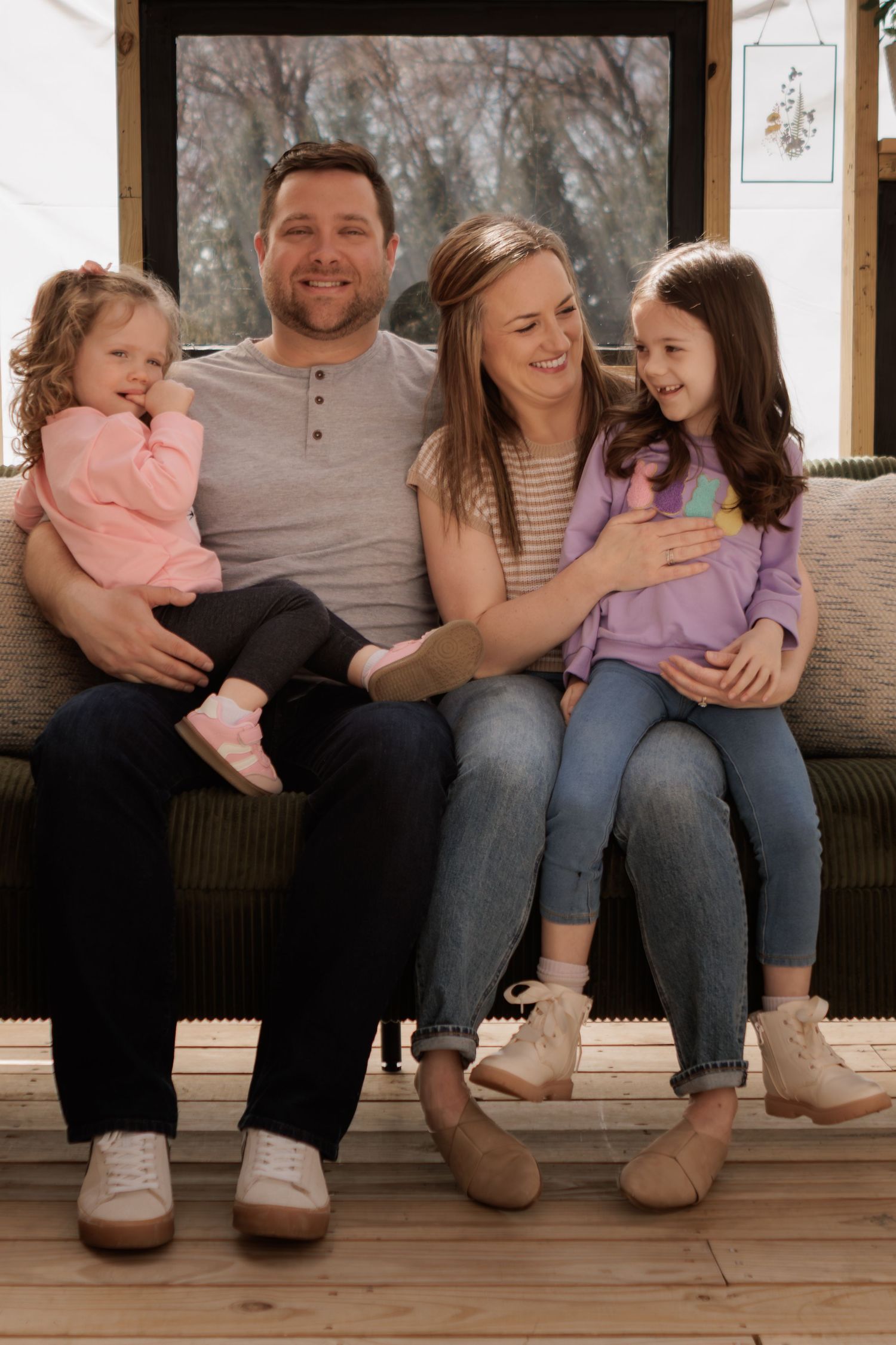 Happy family of four sitting on porch steps, parents smiling with two young daughters in casual clothing.