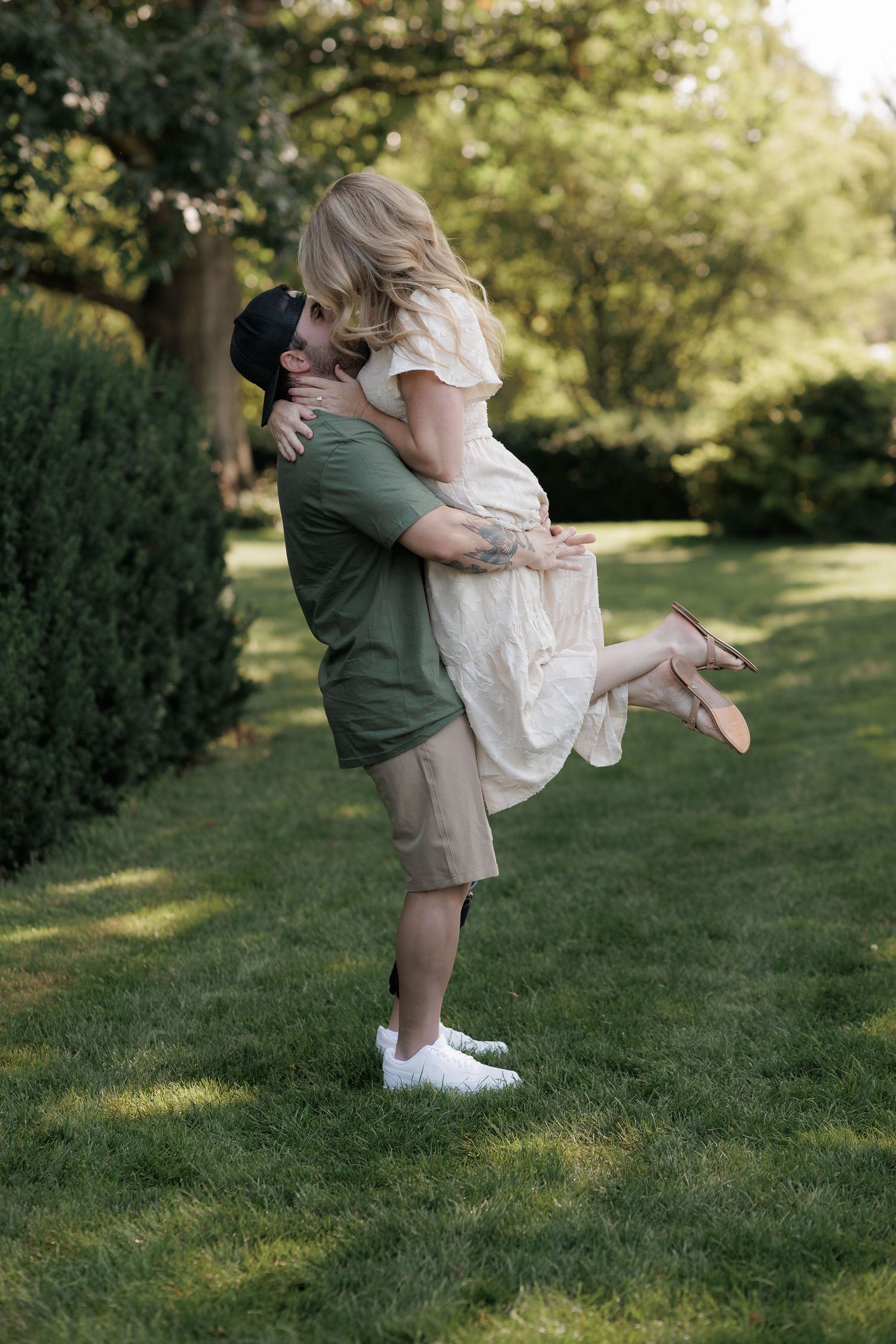 A man lifts a woman in a romantic embrace on a lush green lawn surrounded by trees.