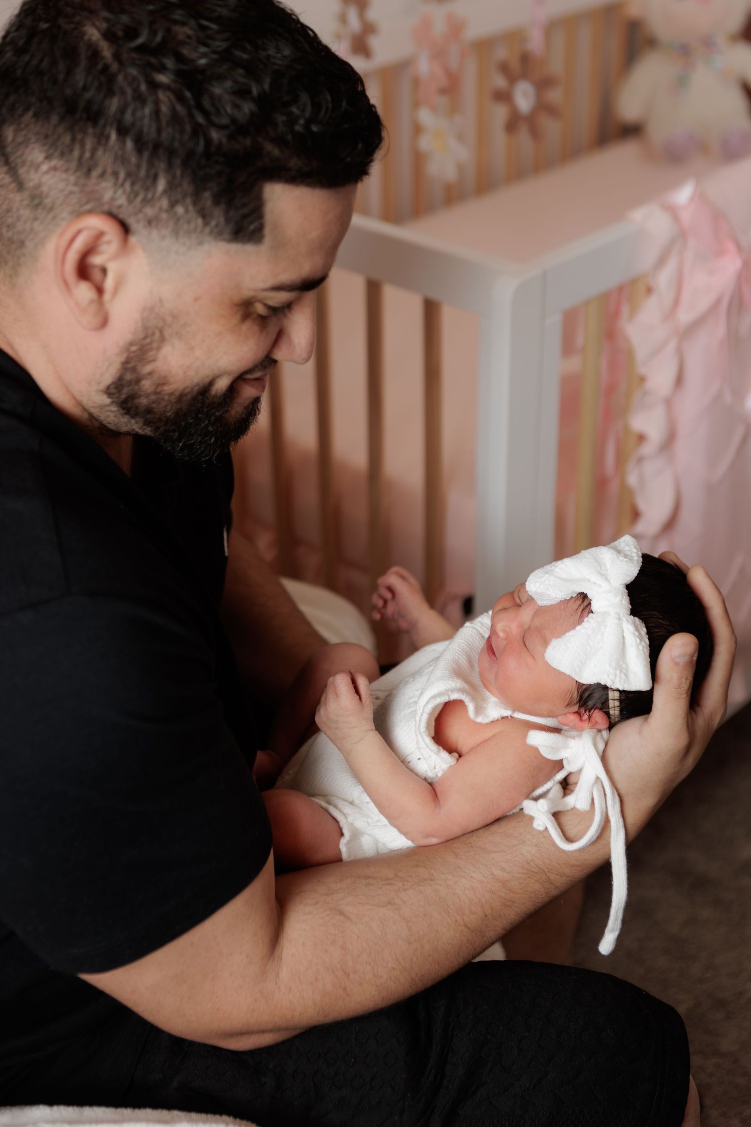 A bearded man in a black shirt gently cradles a sleeping newborn girl wearing a white bow headband beside a crib in a pink nursery.