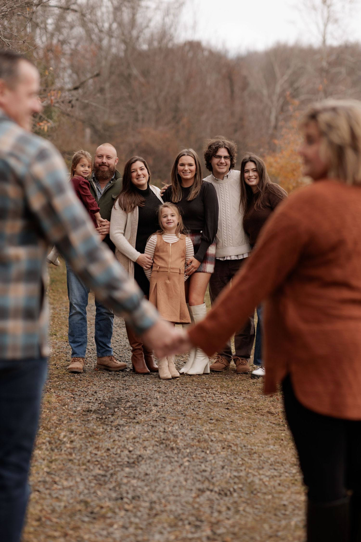 Large family gathering outdoors in autumn, holding hands on a gravel path surrounded by bare trees.
