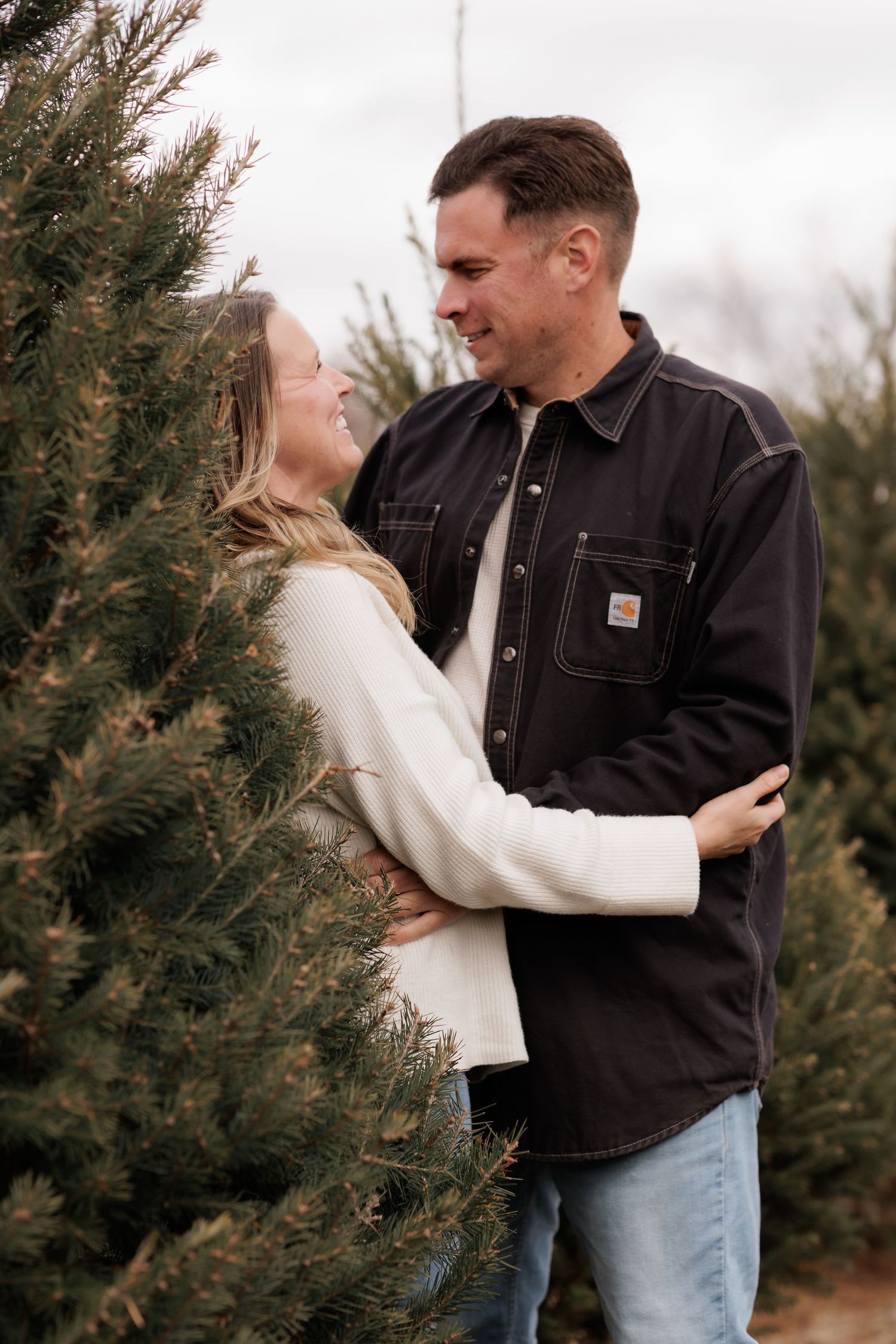 Couple embracing and laughing together among Christmas trees.