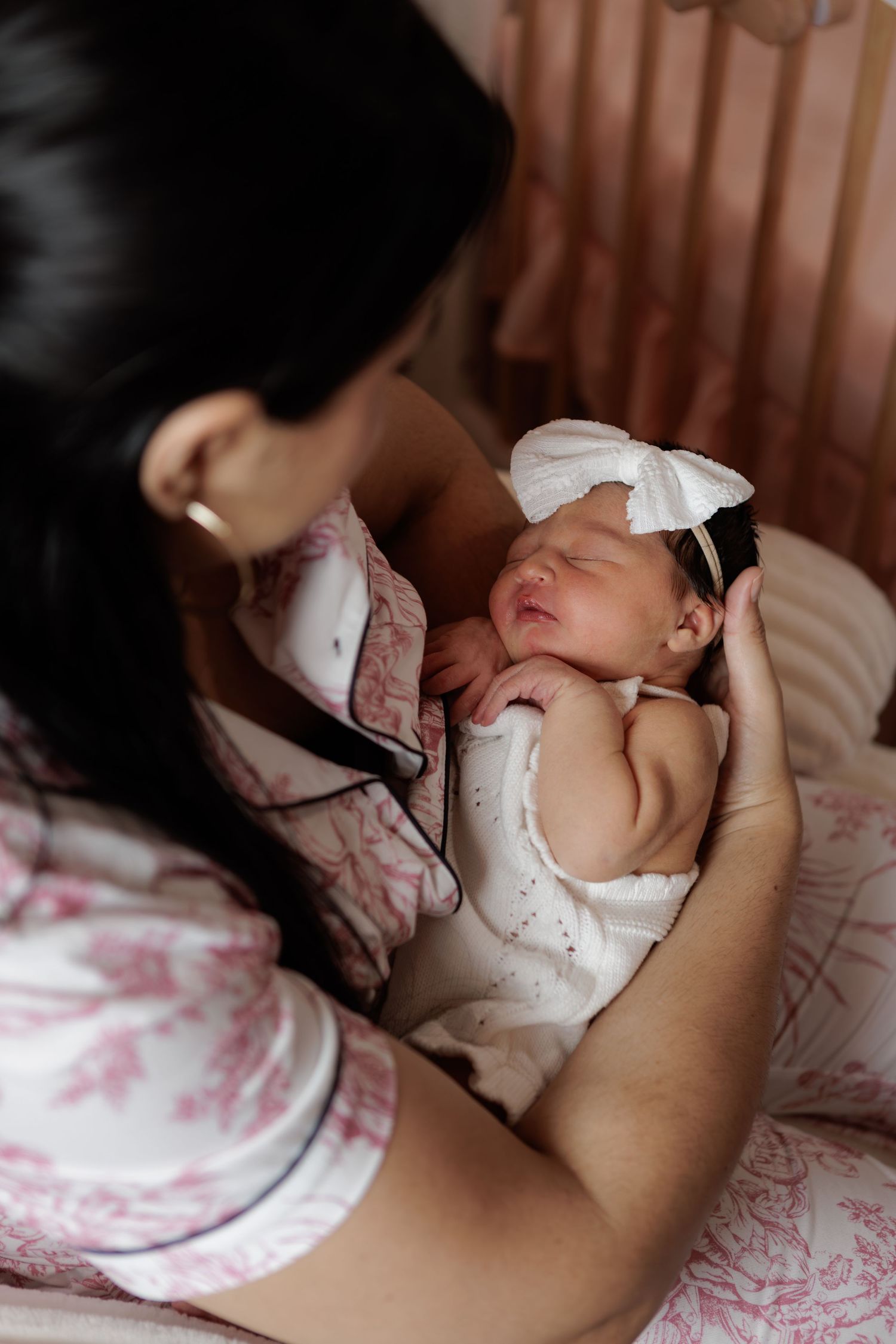 Mother in a pink floral robe cradles a sleeping newborn wearing a white bow headband beside a wooden crib.