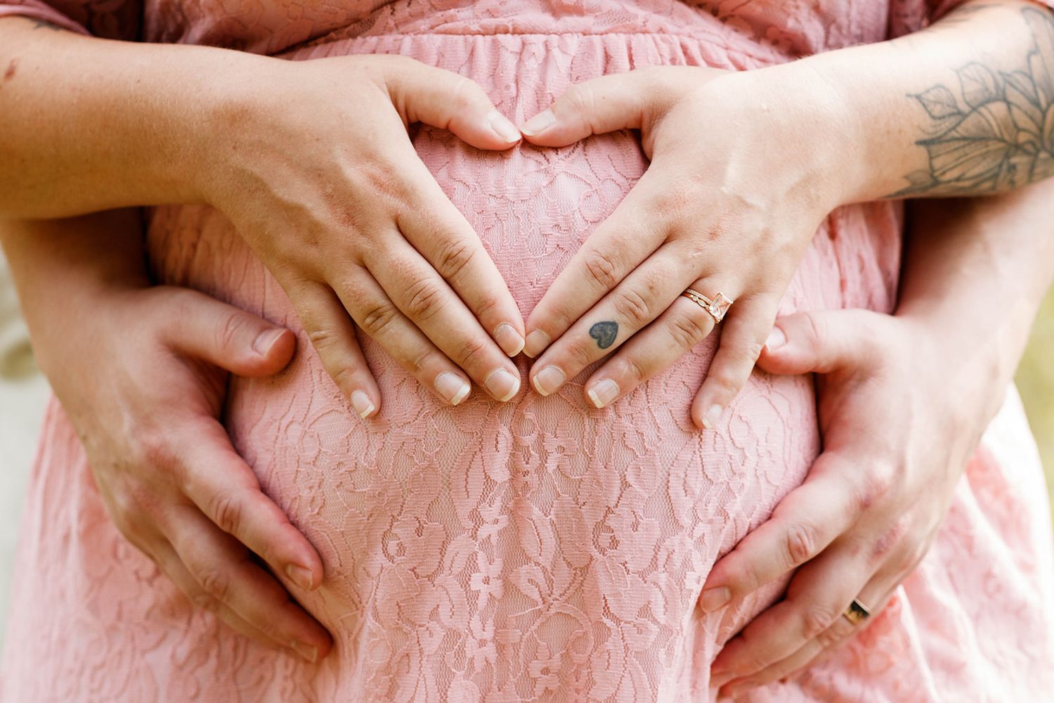 Two pairs of hands forming a heart shape on a pregnant woman's baby bump in a pink dress.