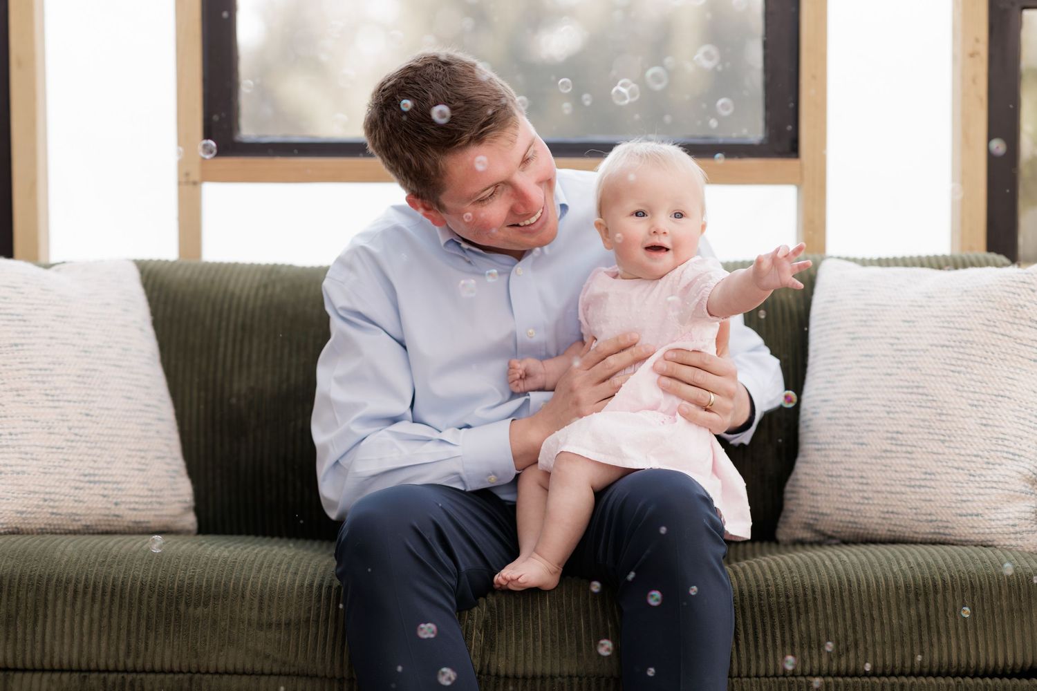 Father sitting on green sofa holding baby girl in pink dress, both smiling as bubbles float around them indoors.