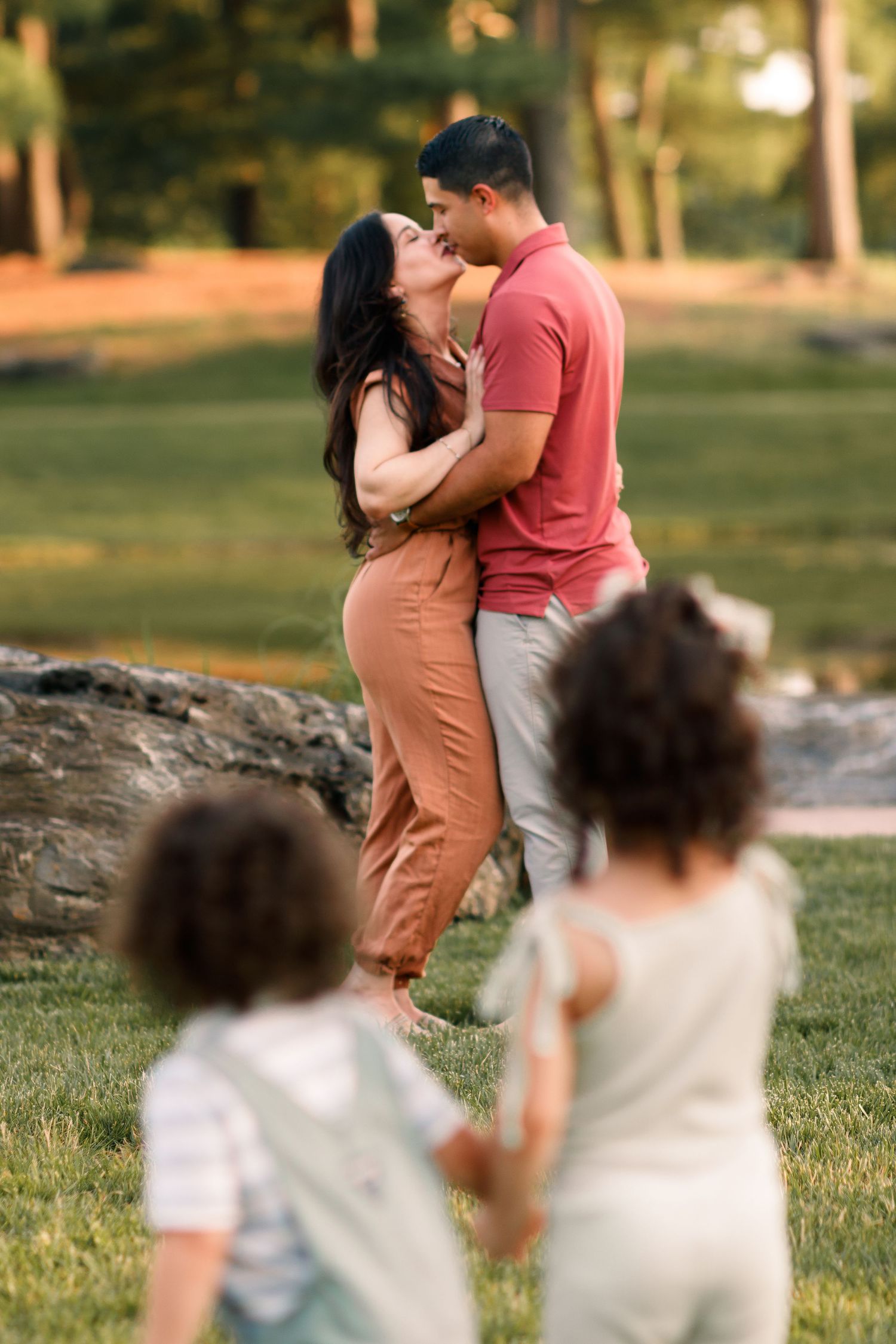 A couple shares a romantic moment in a park while children play in the foreground on a sunny afternoon.