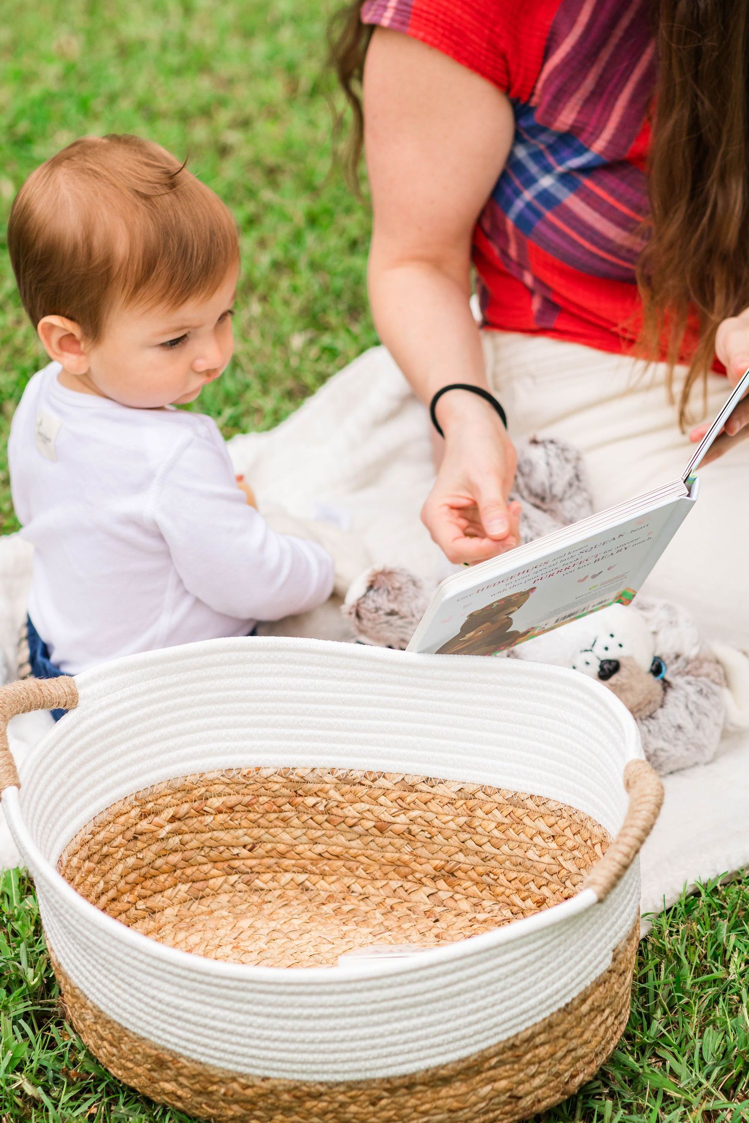 A large woven basket filled with sand or sensory material sits on grass while someone reads a book to a small child.