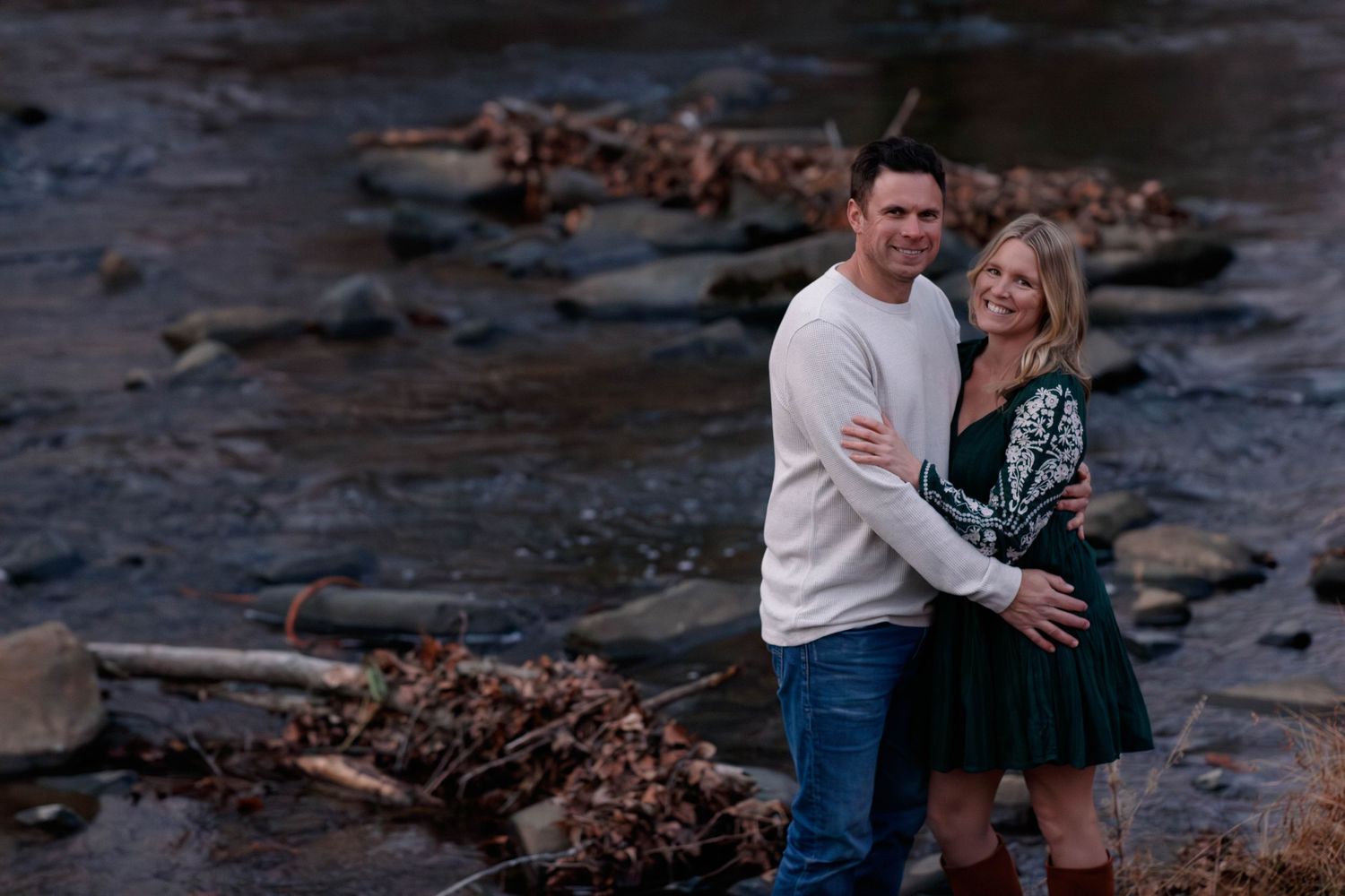 A couple embraces on rocky terrain near the water during an outdoor photoshoot at dusk.